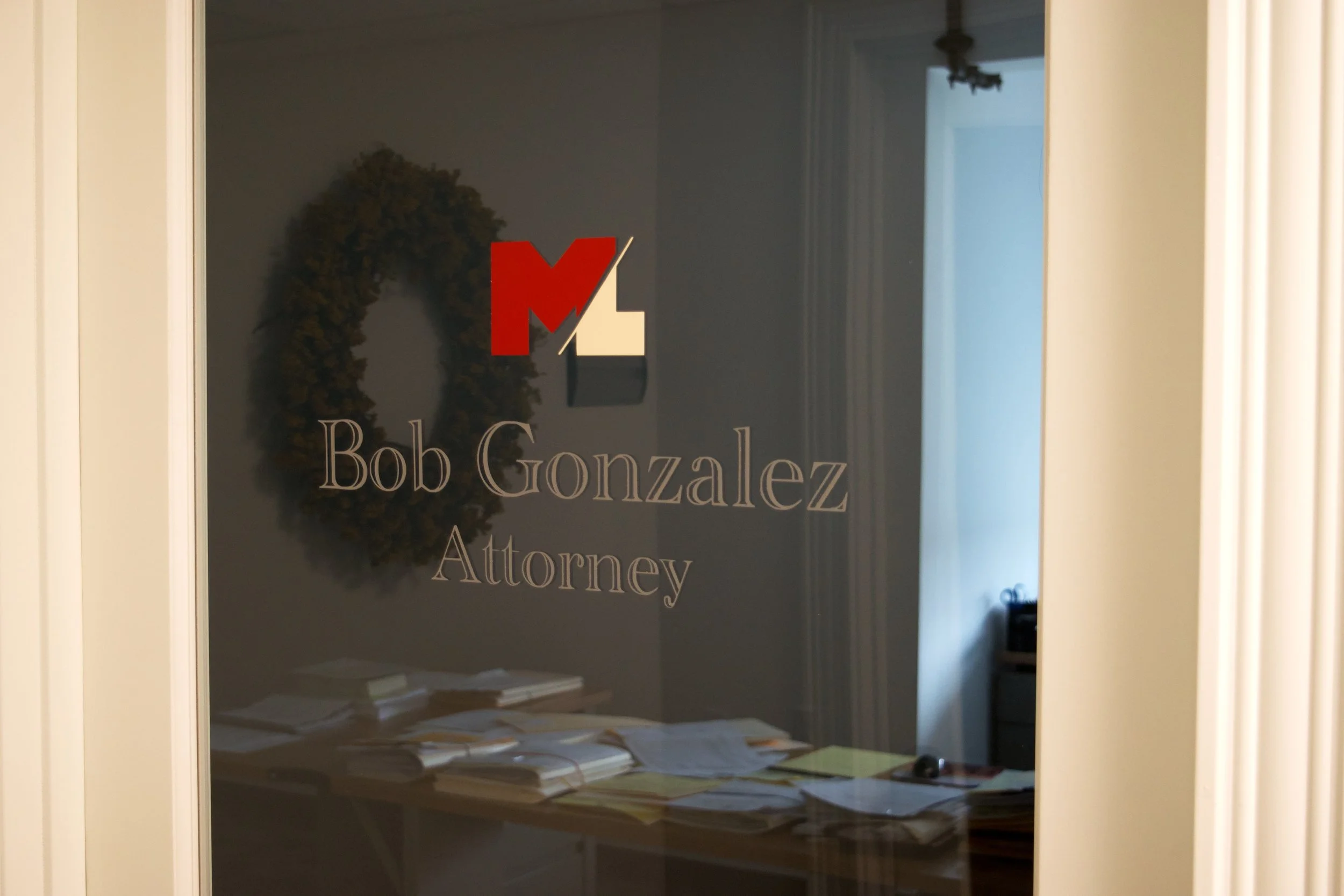 Office door with glass panel displaying name and title of attorney, Bob Gonzalez, with piles of papers and office furniture visible inside.