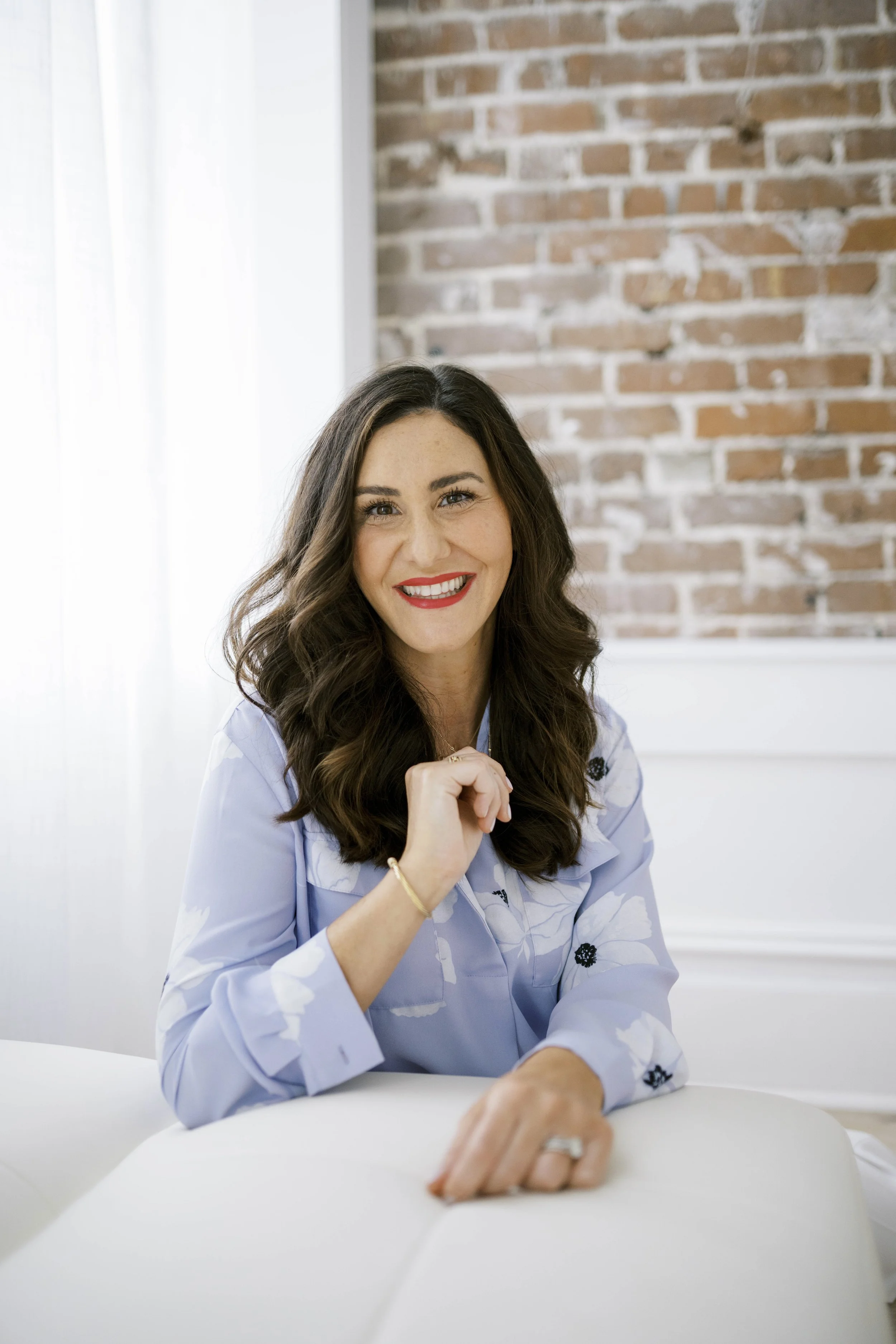 A woman with long dark hair, red lipstick, and a blue floral blouse sitting at a table, smiling at the camera in front of a brick wall.