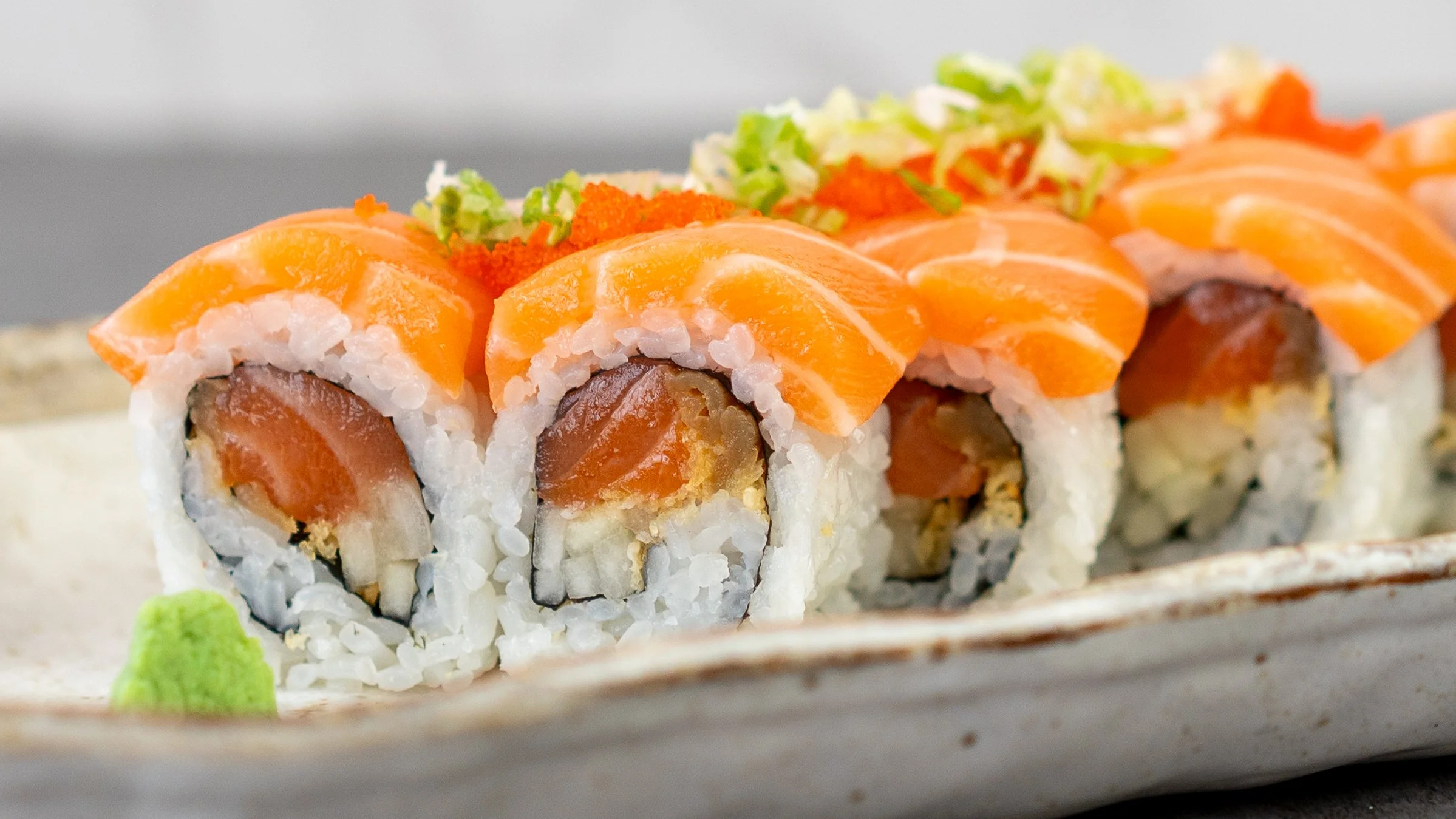 A macro shot of a special roll featuring salmon. The roll comes in 8 pieces, with salmon, cucumber, kampyo, and crunch, rolled with nori and sushi rice, and topped with more salmon, tobiko, and scallions. There is a bit of wasabi in the foreground.