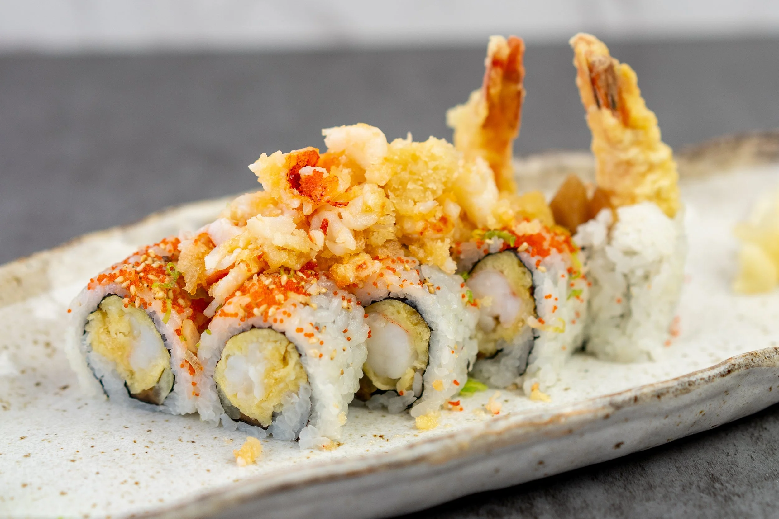 A close up shot of a special roll featuring Lobster. 8 piece maki sushi, with shrimp & lobster tempura, rolled in nori and sushi rice, topped with sesame seeds, tobiko, and torched lobster bits. Shrimp tails still up at the end of the roll.
