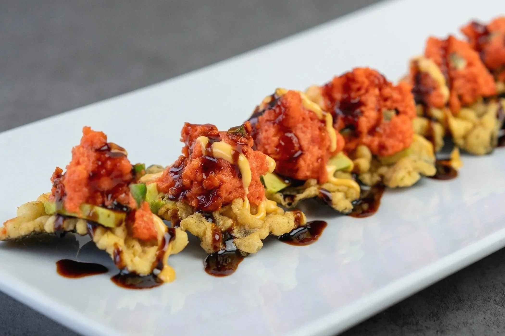 A close up macro shot of a sushi bar appetizer created and named for a customer named David. It features wasabi seaweed tempura chips, laid out in a line, topped with chopped spicy mayo, avocado bits, eel sauce, and spicy mayo.