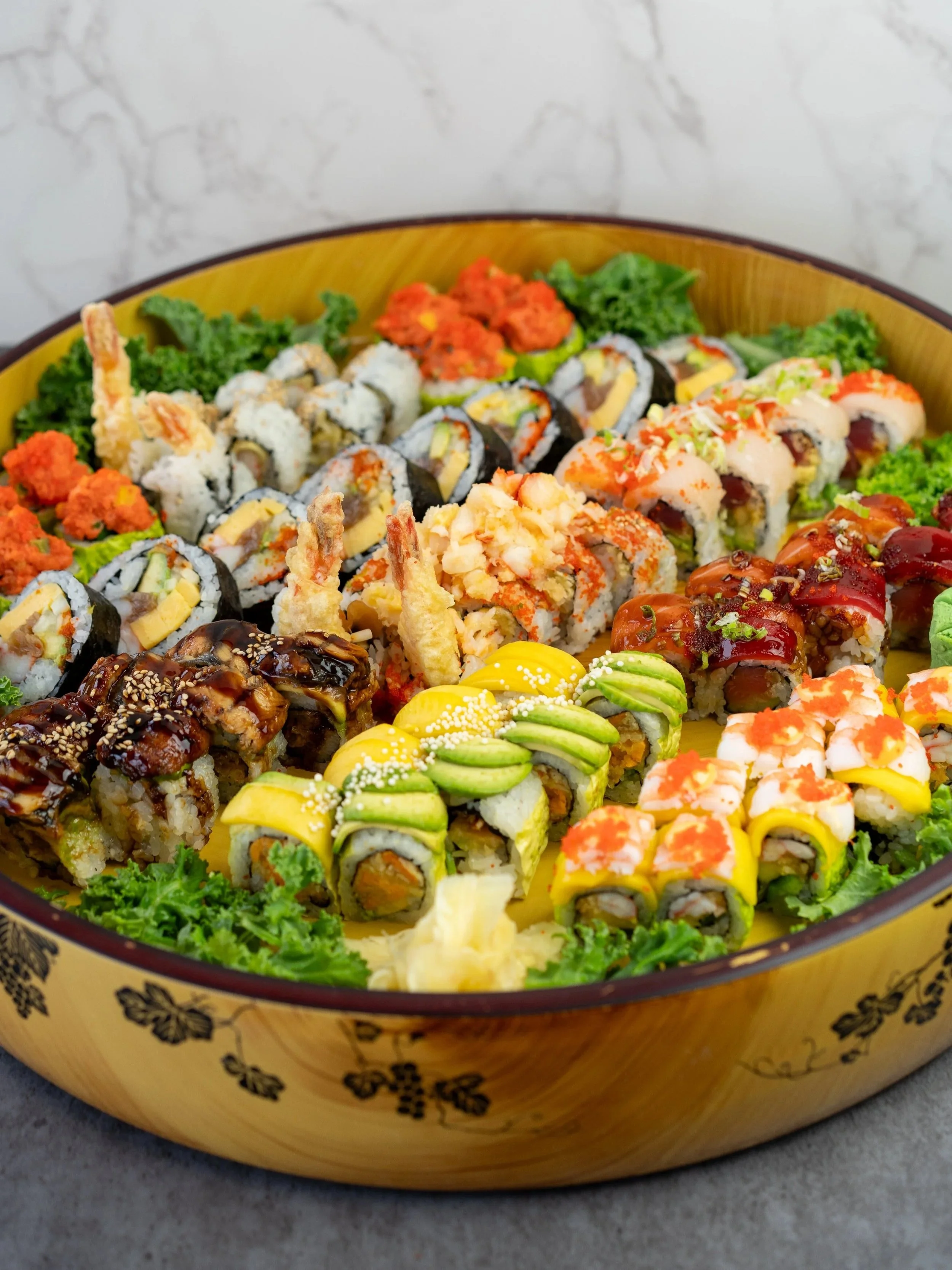 A wide shot of a sushi platter with various rolls containing tuna, salmon, crab, shrimp, lobster, eel, mango, avocado, and more. Garnished with scallions, kale, tobiko, and more.