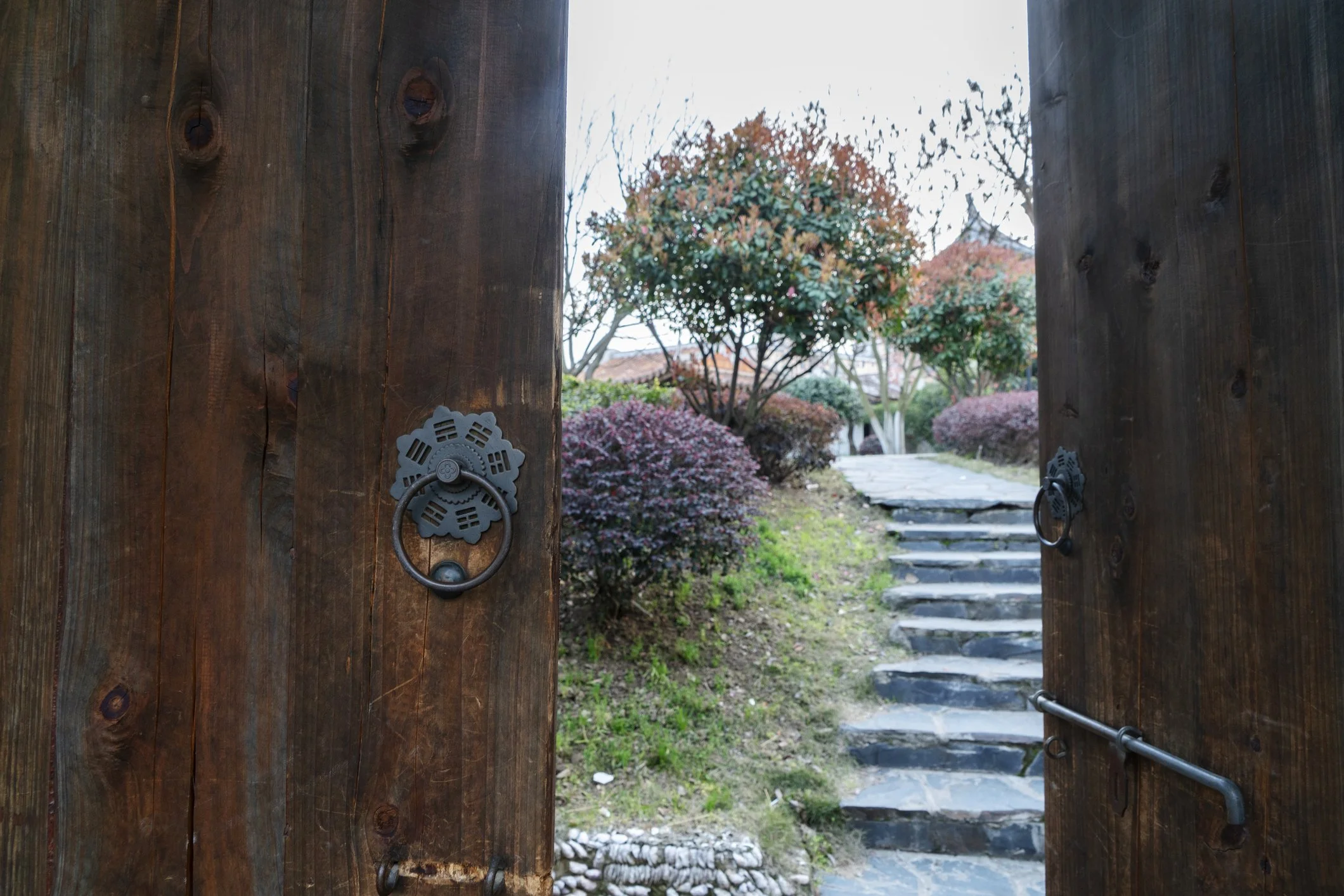 Wooden gate partially open revealing a garden with trees and shrubs, stone pathway, and steps in a landscape.