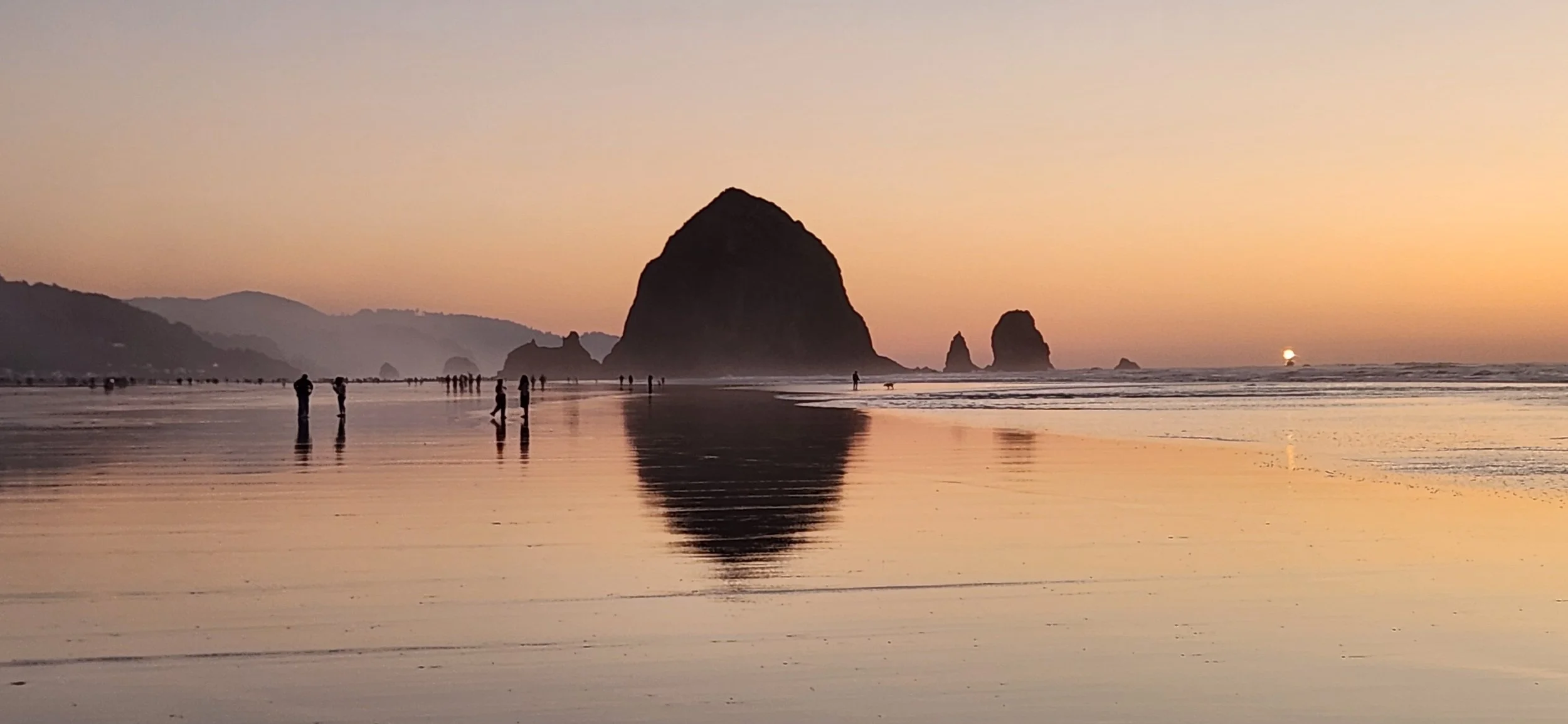 Sunset at a beach with large rock formations, people walking along the shore, and reflections on the wet sand.