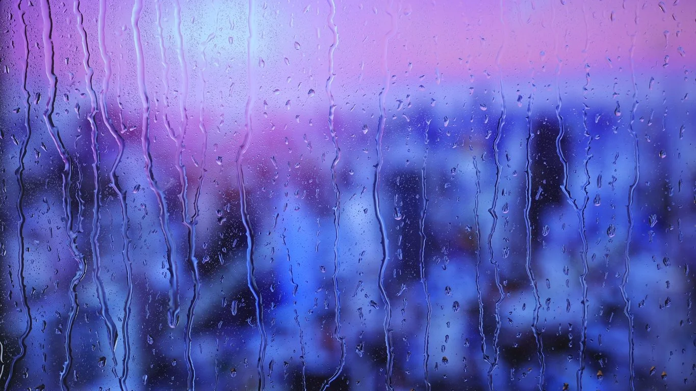 Rain streaks on a window with a blurred sunset or sunrise in the background, showing shades of purple, pink, and blue.