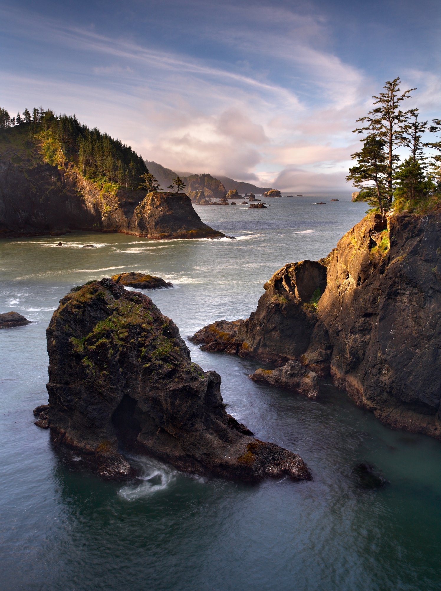 Coastal landscape with rocky cliffs, green trees, and ocean waves under a partly cloudy sky.