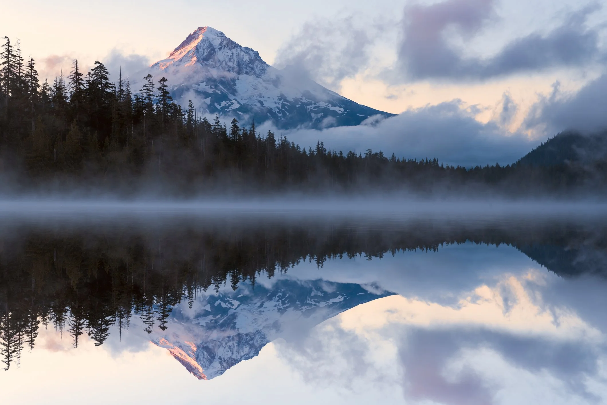 Snow-capped mountain behind a forest with tall trees, reflected in a calm lake with mist at the water's edge.