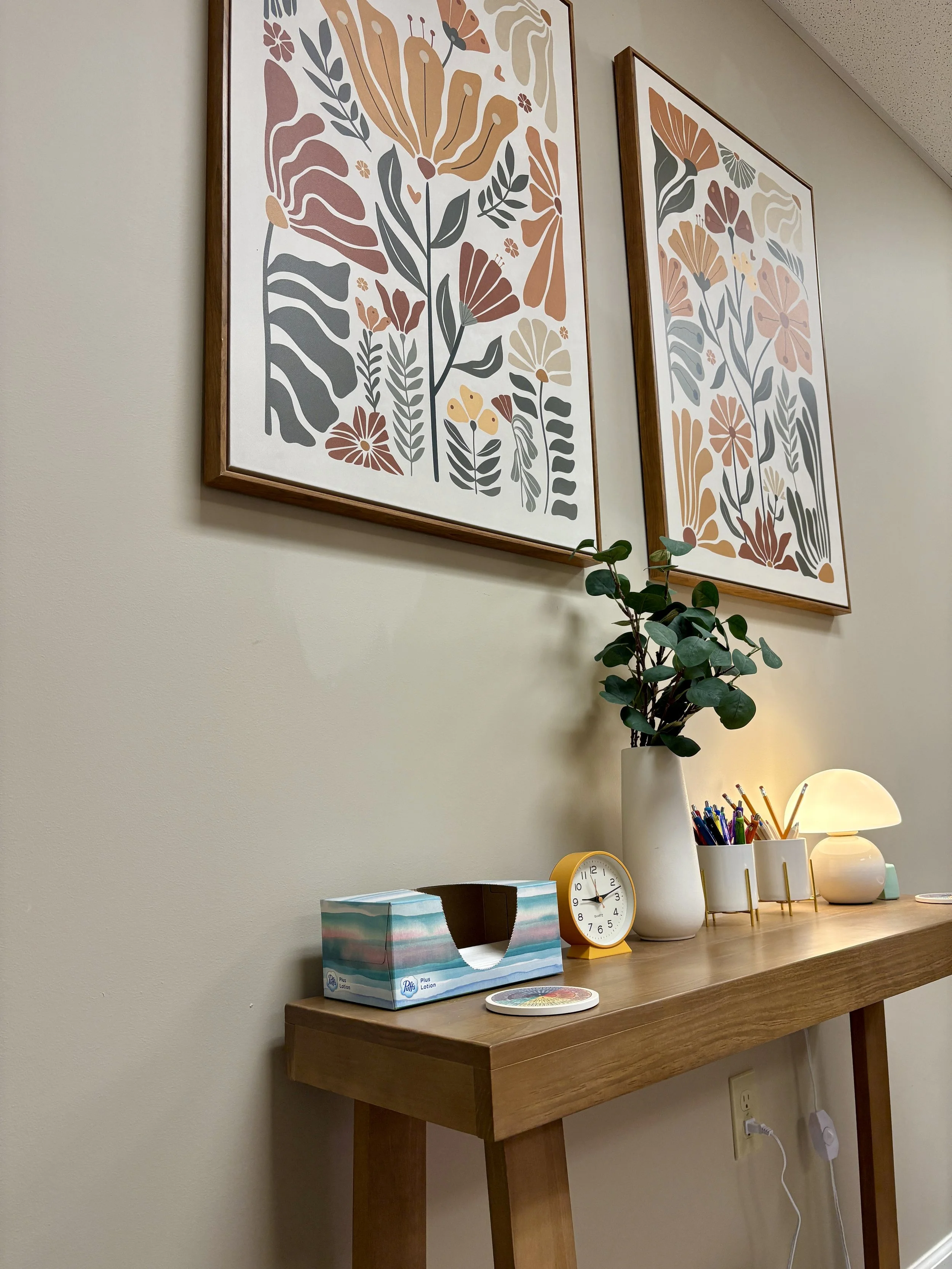 A view from the loveseat of the floral artwork adorning the walls of Fletcher Marron's office for counseling and therapy. There is also tissues, a small orange clock, a vase with eucalyptus, and a mushroom shaped lamp.
