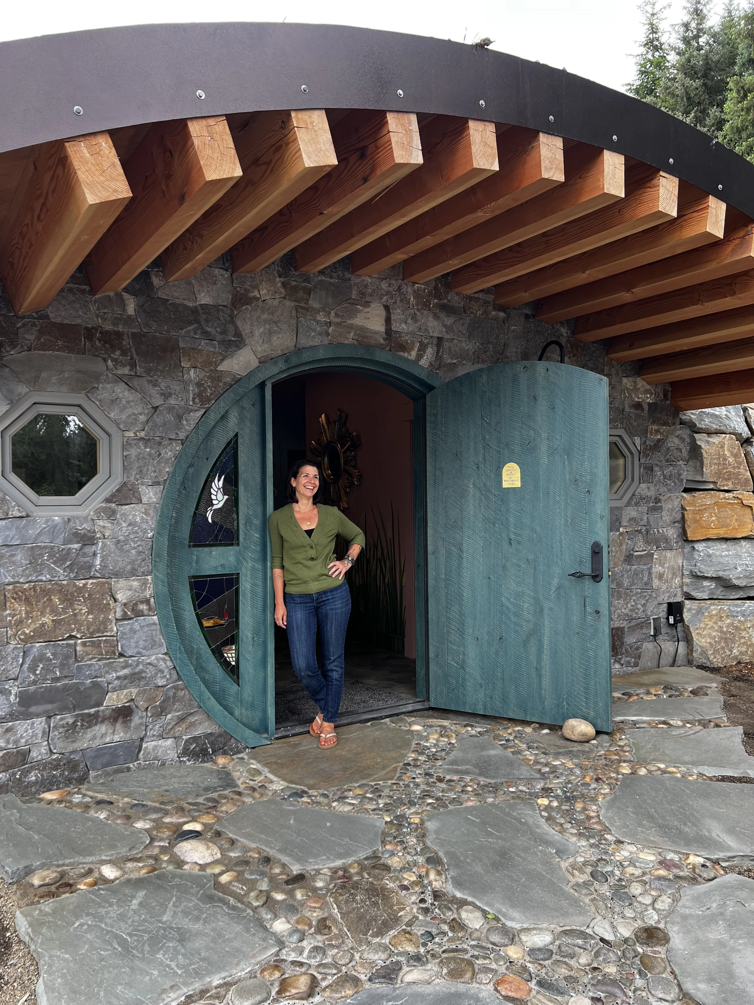 A smiling woman, Cheri Tilford, with dark hair wearing a green cardigan and jeans standing in front of an open green door with round windows, part of a stone hobbit house building, called the Healing Hollow, with a wooden overhang.