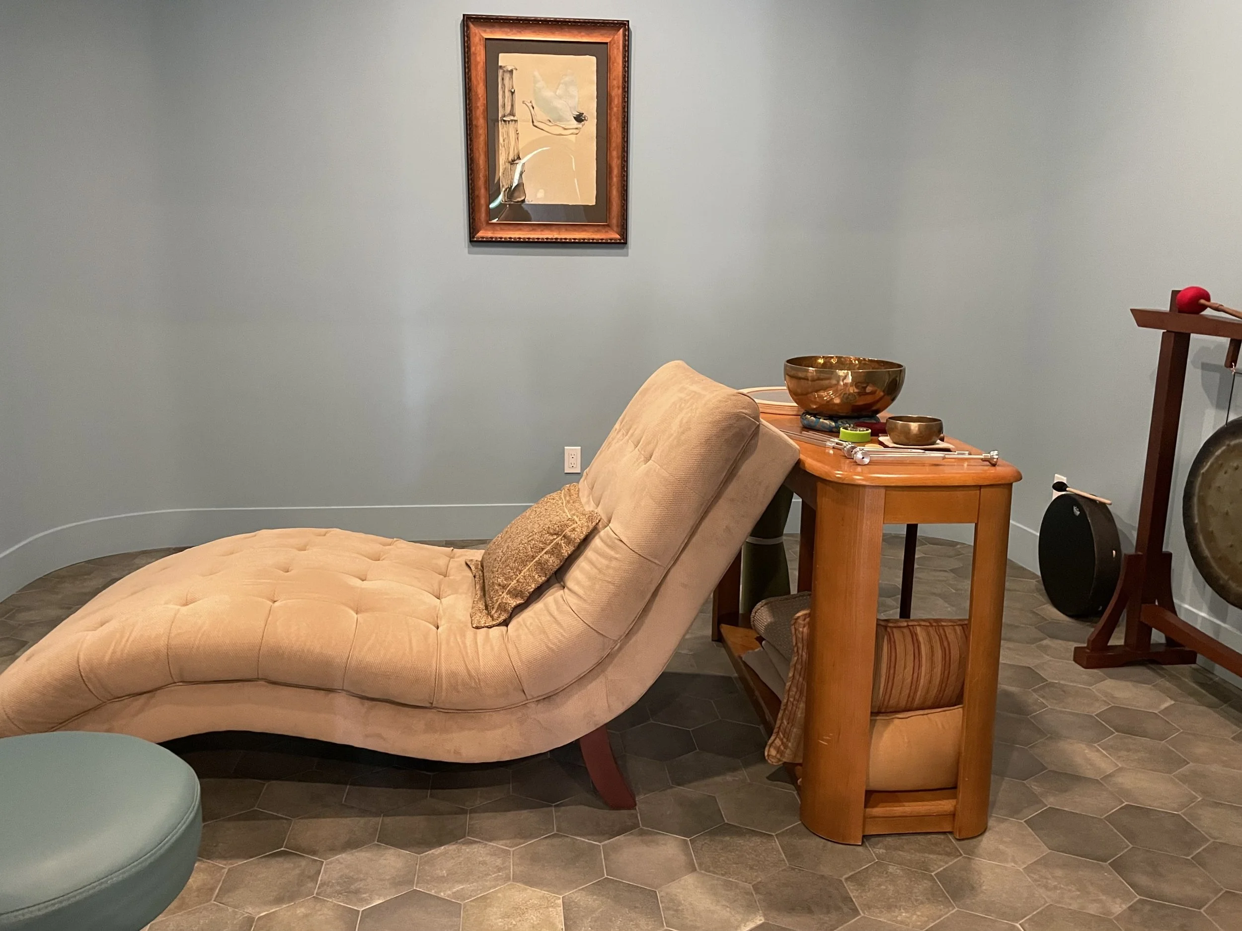 The treatment room of the Healing Hollow with a beige chaise lounge, a wooden side table with bowls and items on it, a meditation gong, and a framed artwork on a blue wall.