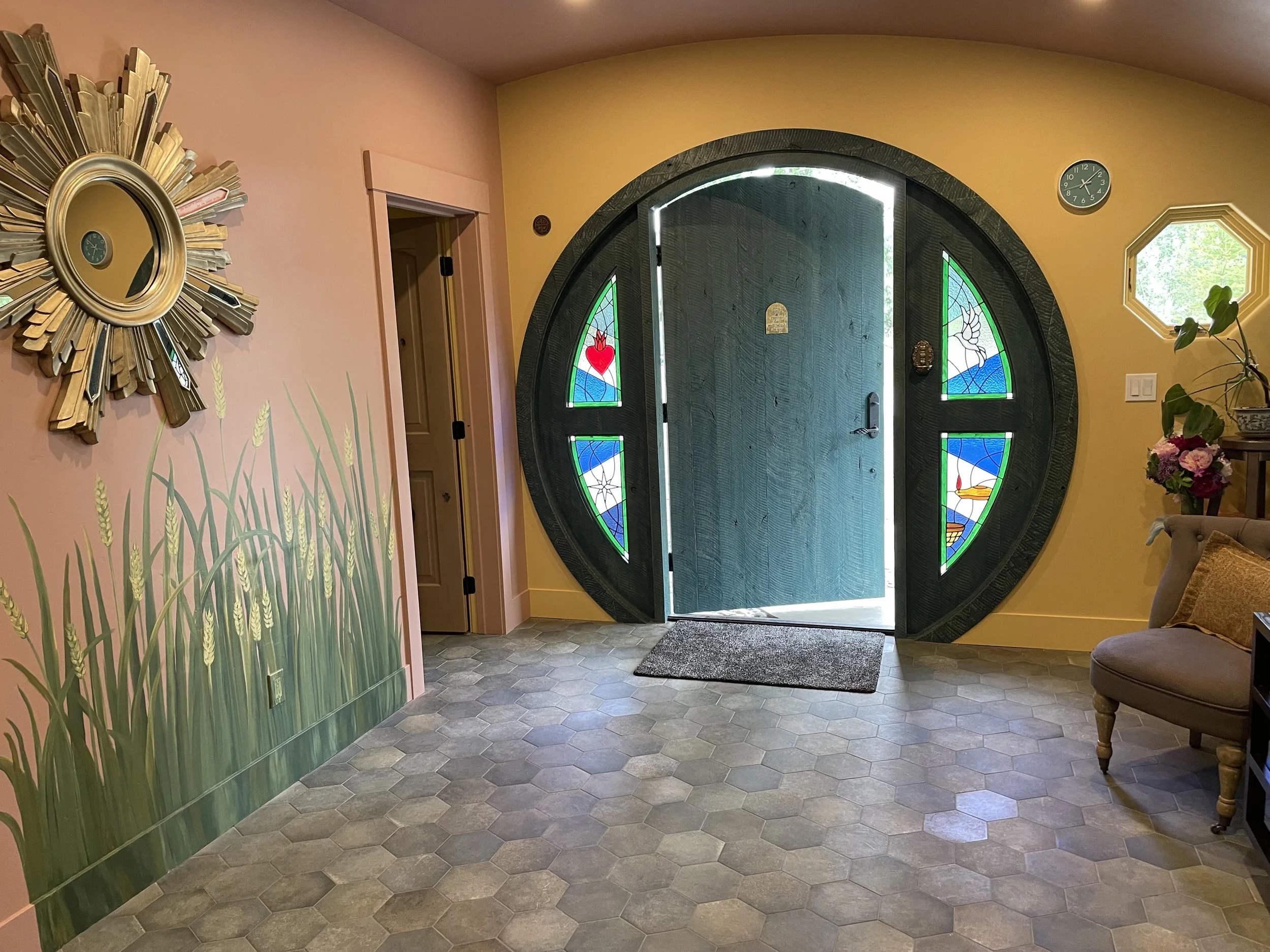 Entryway of the Healing Hollow with a circular door featuring stained glass panels with colorful designs, a stone floor, yellow walls, a floral arrangement, and a mirror with a sunburst frame.