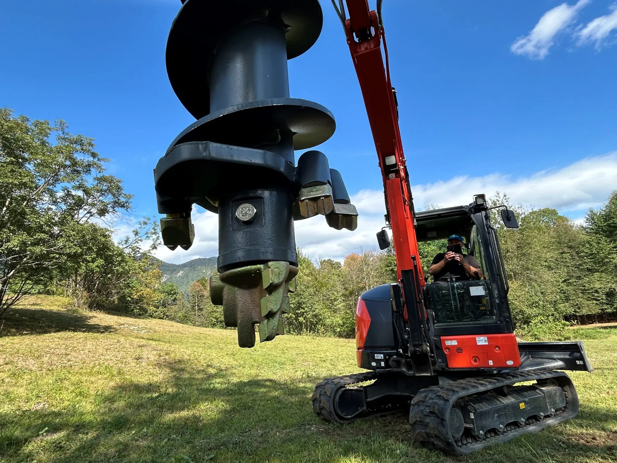 A person operating a small tracked excavator during construction work in a grassy outdoor area with trees and mountains in the background under a blue sky.