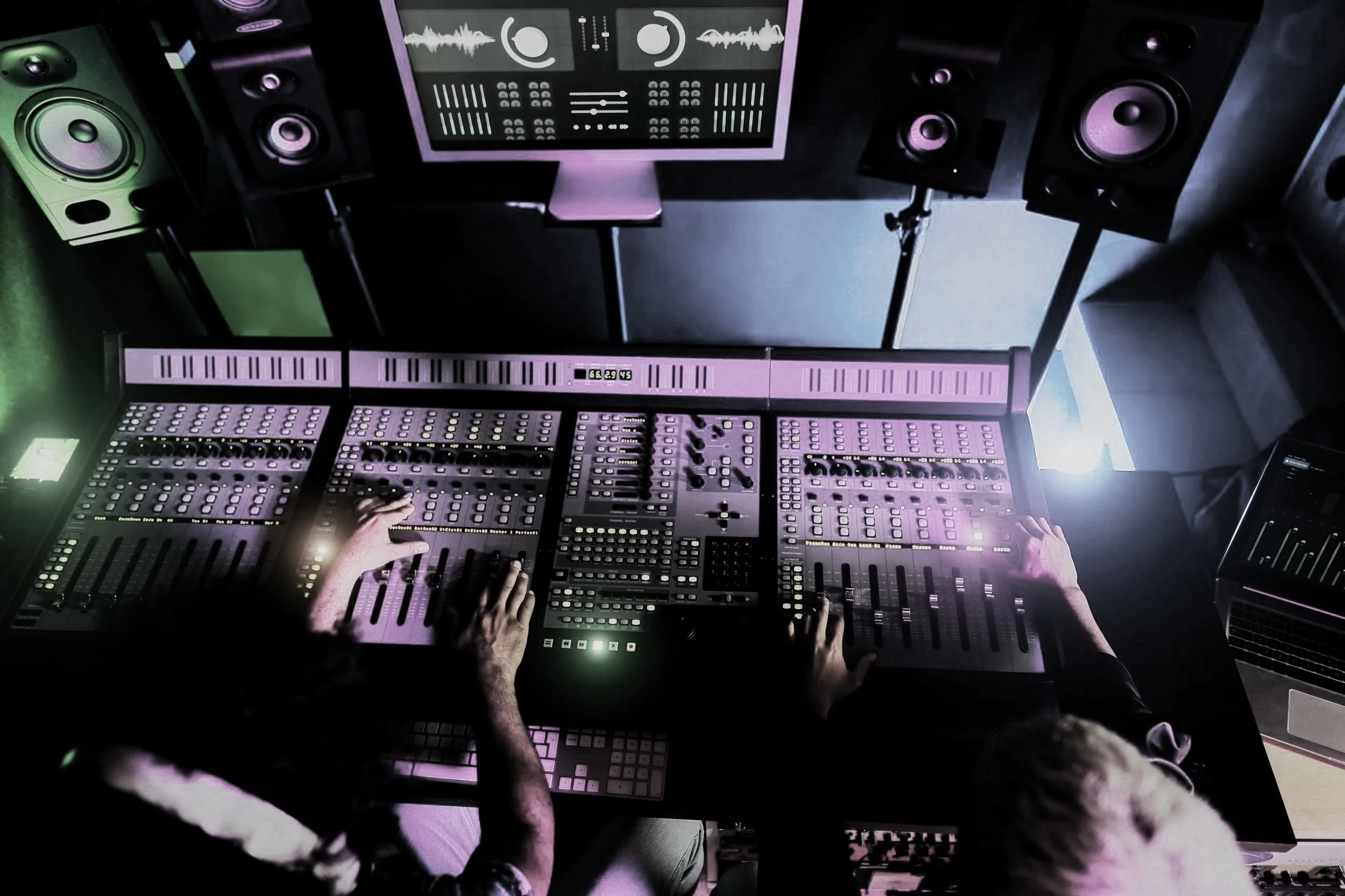 A DJ is operating a large mixing console with multiple sliders and knobs in a dimly lit studio, surrounded by speakers and a computer screen displaying audio waveforms and controls.