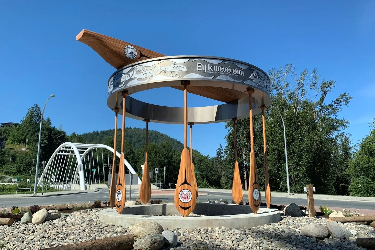 A monument designed as a giant wooden spoon with four wooden paddles hanging from a circular metallic structure. The top of the monument features a large wooden spoon handle, and the circular metallic band has tribal-like engravings and the words 'Eykwe emi.' The monument is placed on a circular concrete base surrounded by rocks and is set outdoors with trees, a bridge, and a highway in the background.