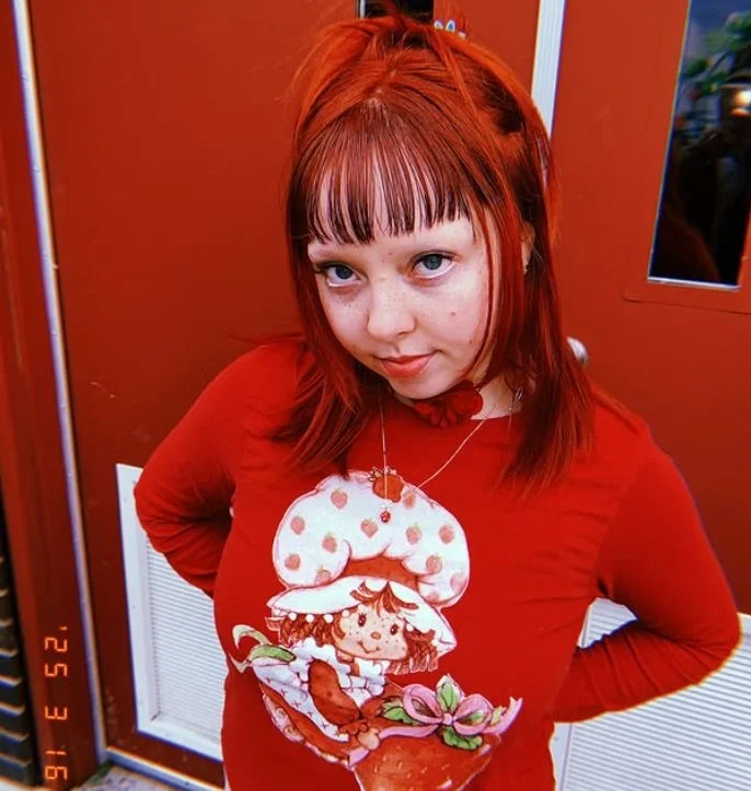 Young woman with red hair and bangs wearing a red shirt and cookie monster necklace posing indoors in front of a red door.