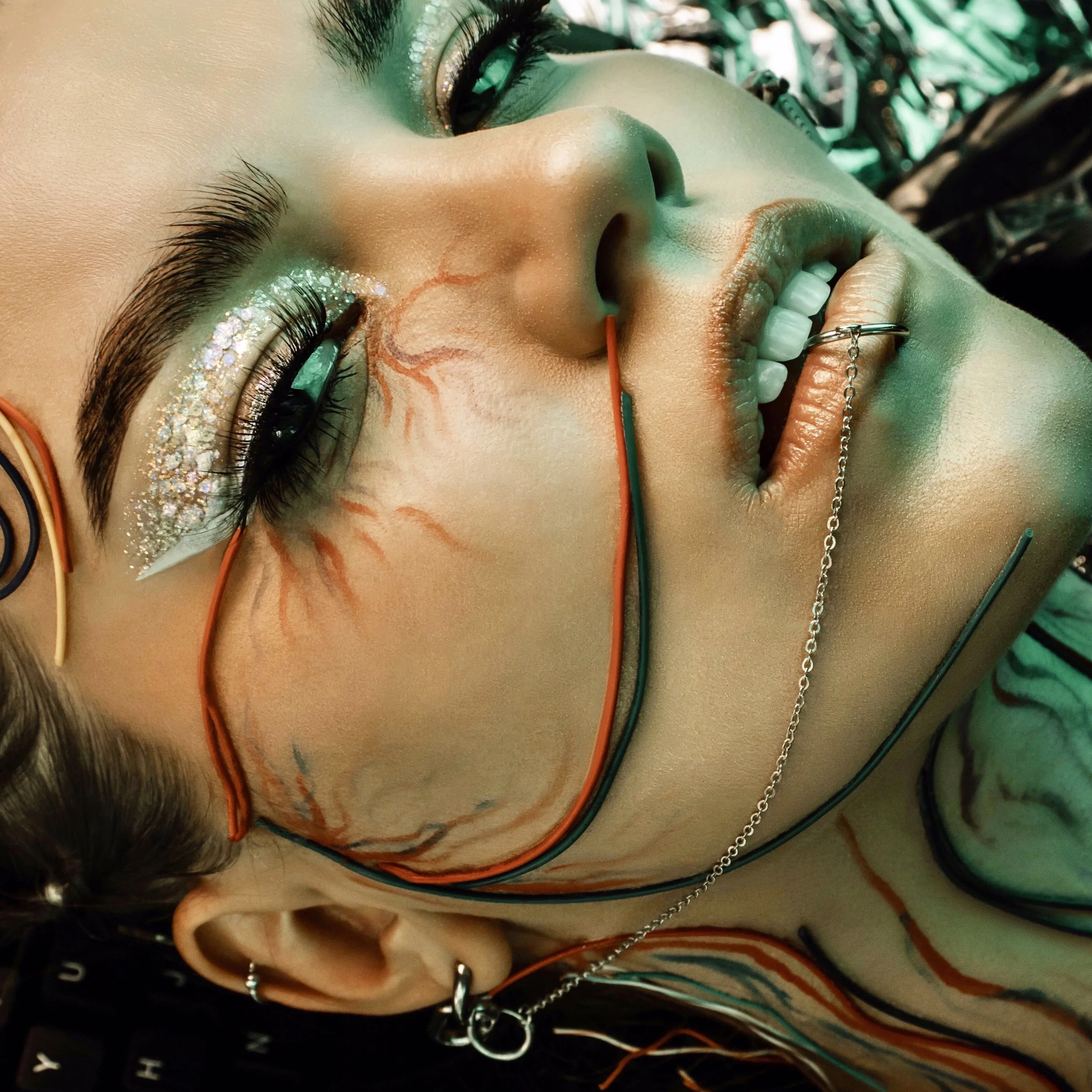 Close-up of a person with artistic makeup, including glitter and colorful paint, lying down with wires and chains connected to their face and head. This is a photo of the founder of No Labels Studio, Maddi Krulicki.