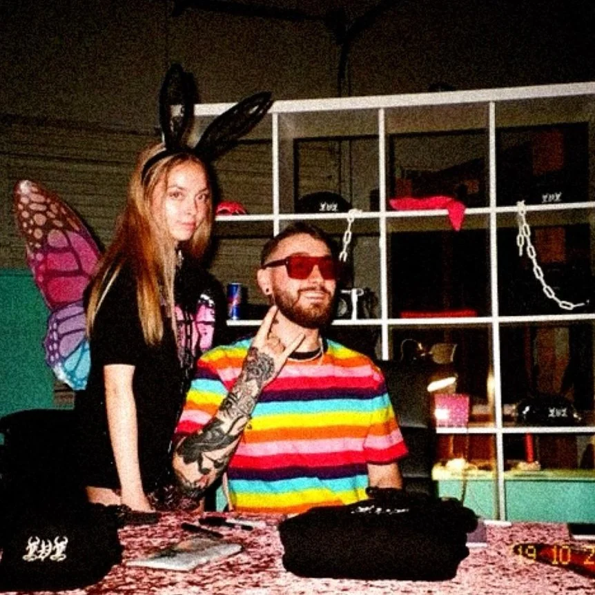 A woman dressed as a butterfly with butterfly wings and bunny ears is standing next to a man wearing colorful striped shirt and red sunglasses, making a peace sign. They are indoors with shelves, chains, and various items in the background, and a table with cloth in front of them.