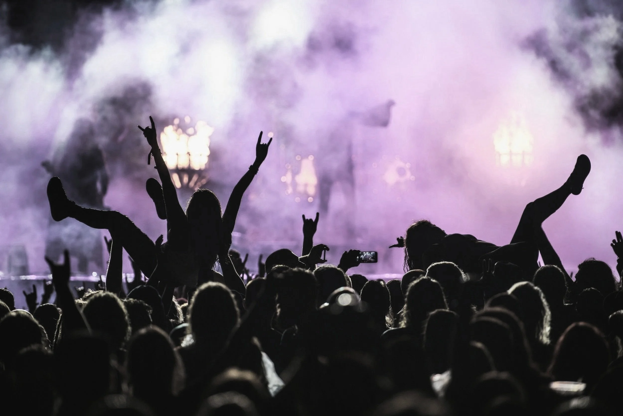 Crowd at a concert with two children lifted on shoulders, silhouetted against a smoky stage with purple lighting.