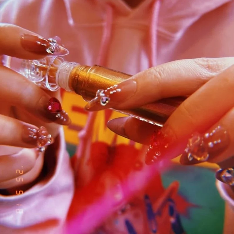 A person with nails decorated with small rhinestones is holding a small vial of liquid and a golden applicator, with colorful clothing in the background.