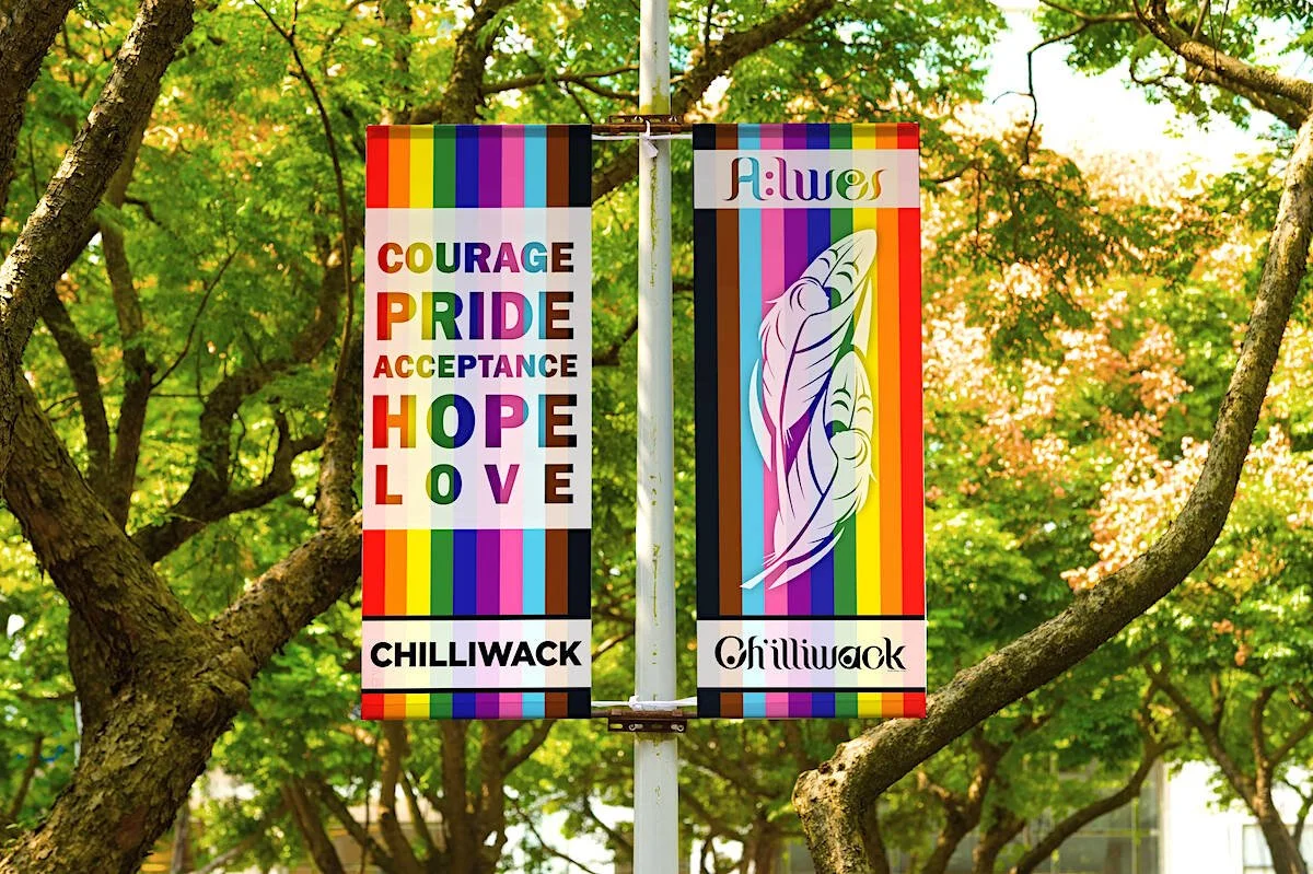 Two banners hanging from a pole between trees, featuring rainbow colors and messages of LGBTQ+ pride and acceptance. One banner reads 'Courage, Pride, Acceptance, Hope, Love' and the other says 'Always' with a dove illustration. The location is Chilliwack.
