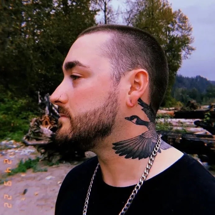 Profile of a young man with a beard, wearing a chain necklace and a black shirt, showcasing a large tattoo of a flying duck on his neck, outdoors near a river with trees in the background.