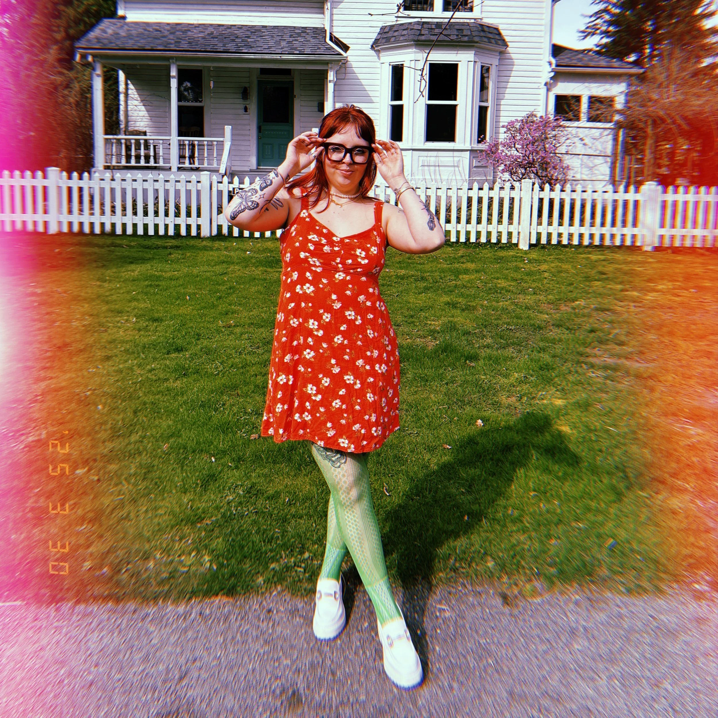 A woman in a red floral dress with green patterned tights and white shoes standing on a sidewalk in front of a white house with a picket fence and a flowering tree.