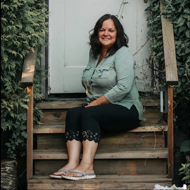 A woman sitting on wooden steps outside next to a white door, surrounded by greenery.