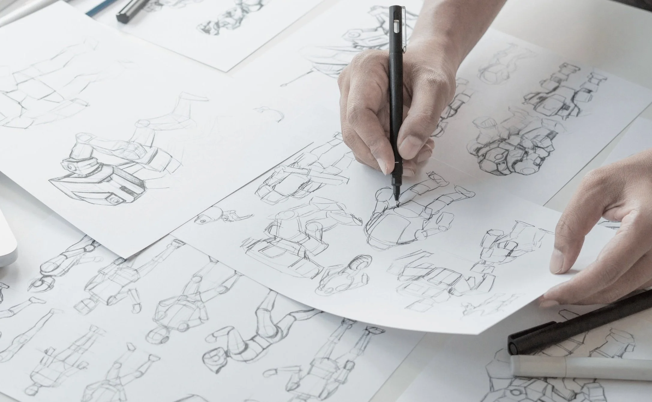 Person sketching robot designs on paper with pencils and pens, surrounded by several sheets of drawings and sketches.
