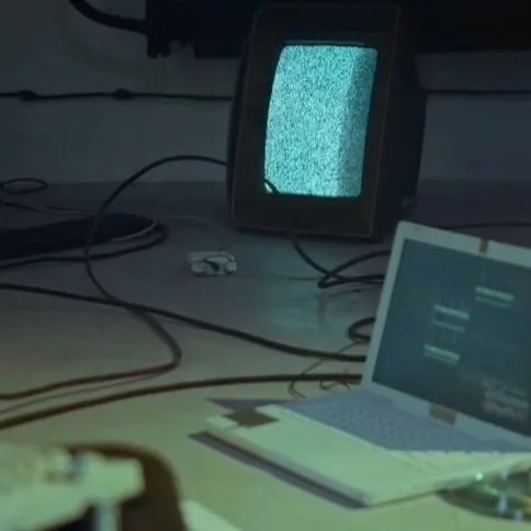 A pixelated television displaying static, a laptop with a dark screen, and various electronic cables on a desk.