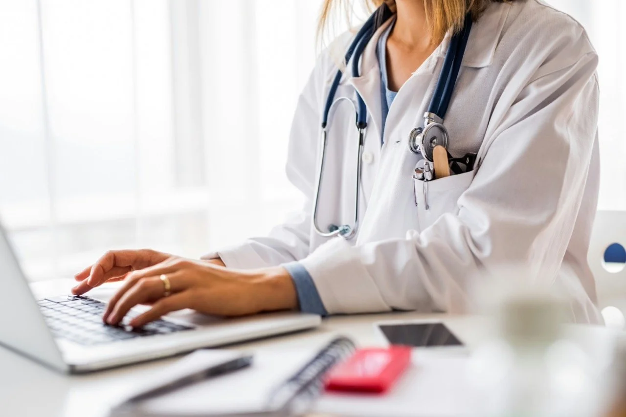 Close-up of a doctor in a white coat with a stethoscope, working on a laptop at a desk in a bright room.