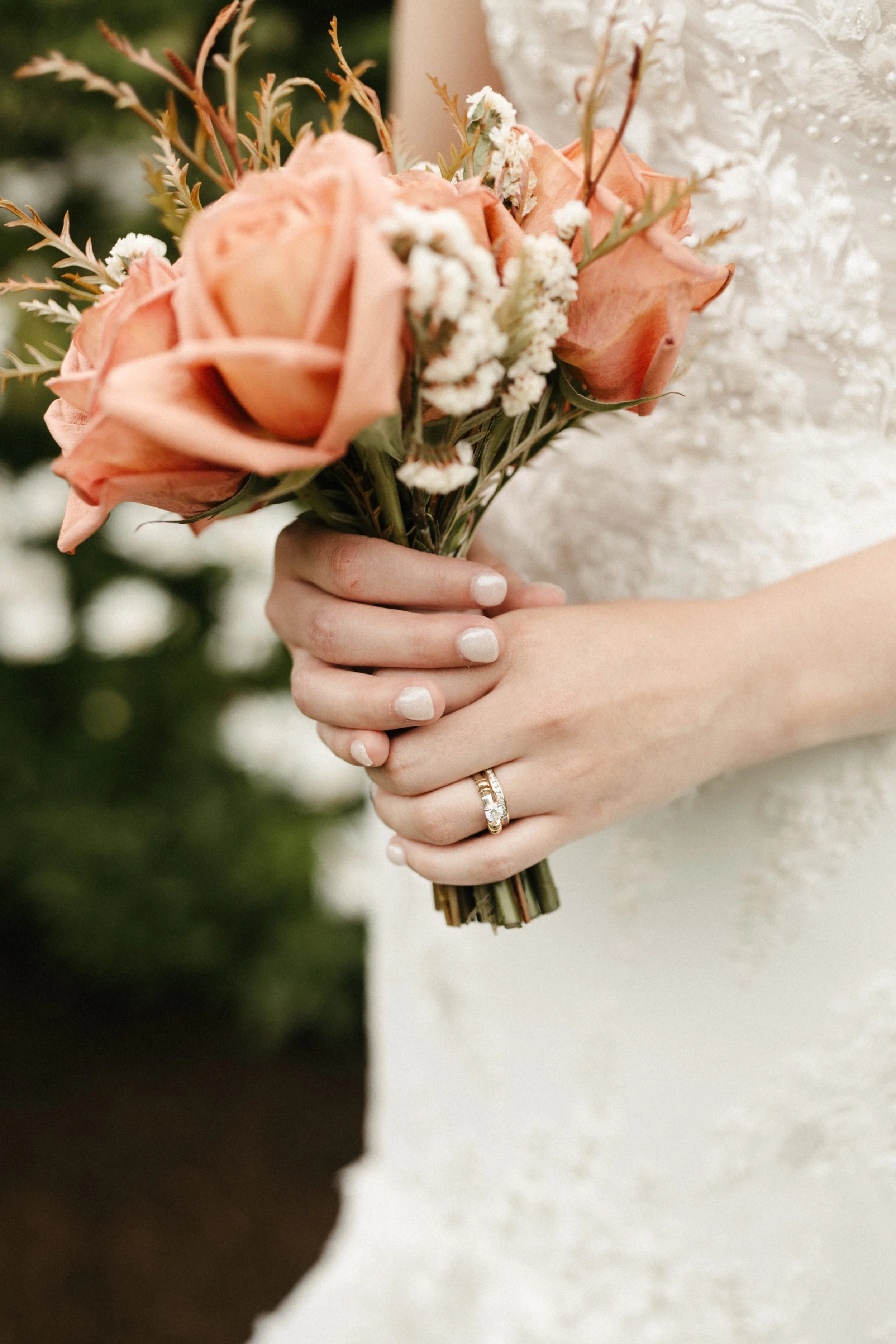 A bride holding a bouquet of peach roses, white flowers, and greenery, with her hands showing a wedding ring.