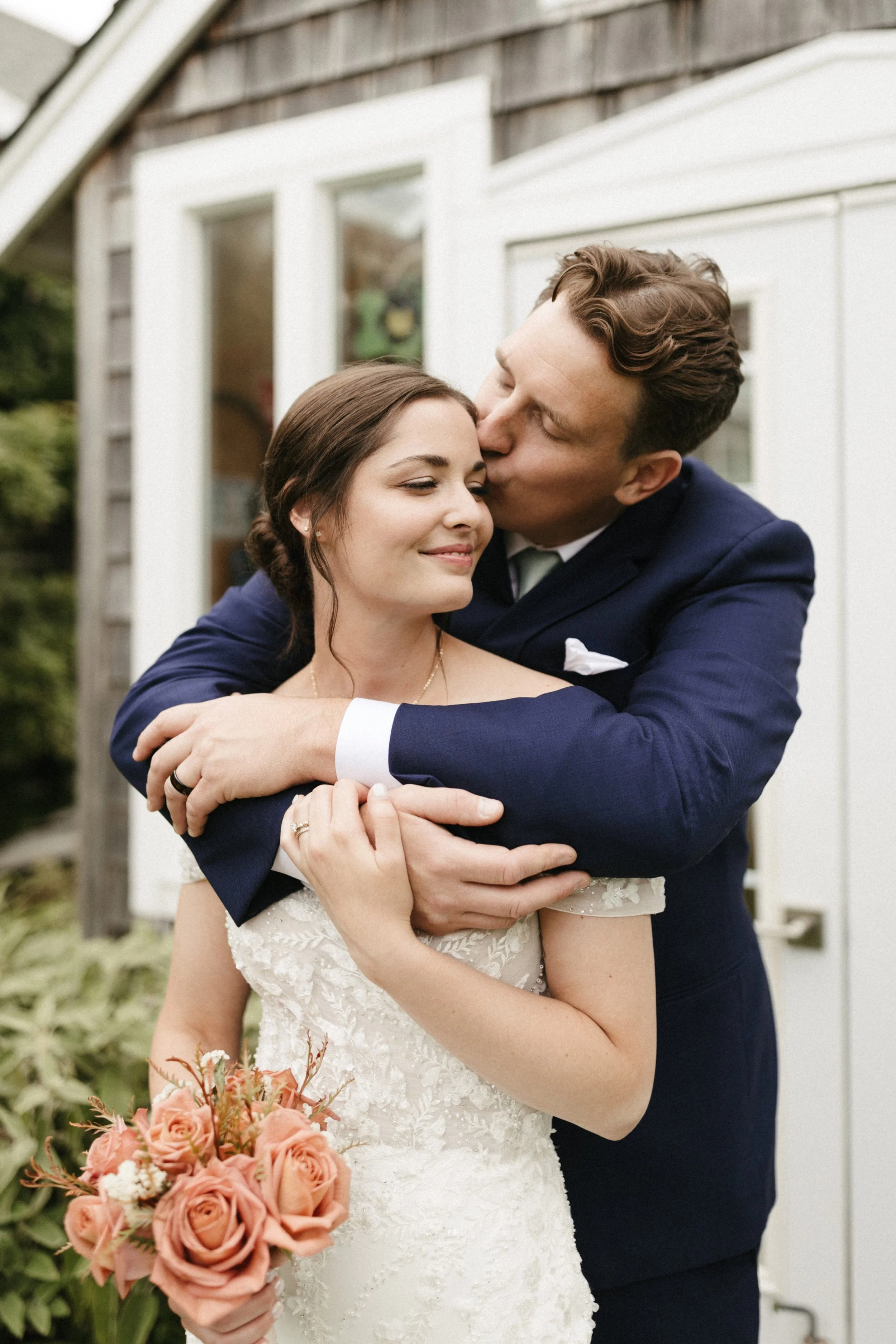 A bride and groom embrace outside a house, with the groom kissing the bride's temple. The bride holds a bouquet of pink roses and wears a white lace wedding dress, while the groom is dressed in a navy suit.