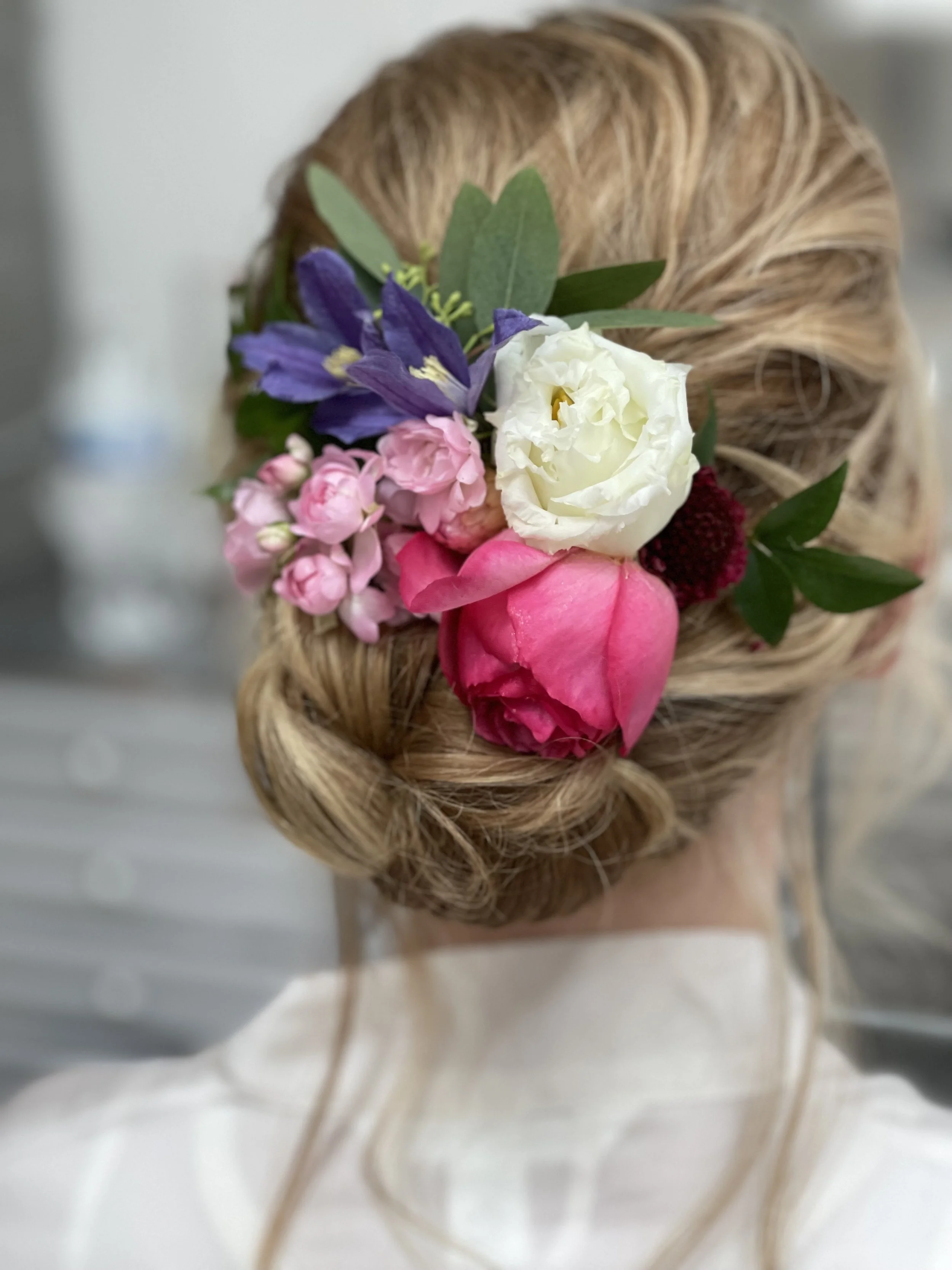 Close-up of a woman's updo hairstyle decorated with a floral arrangement including pink and white roses, purple flowers, and greenery.