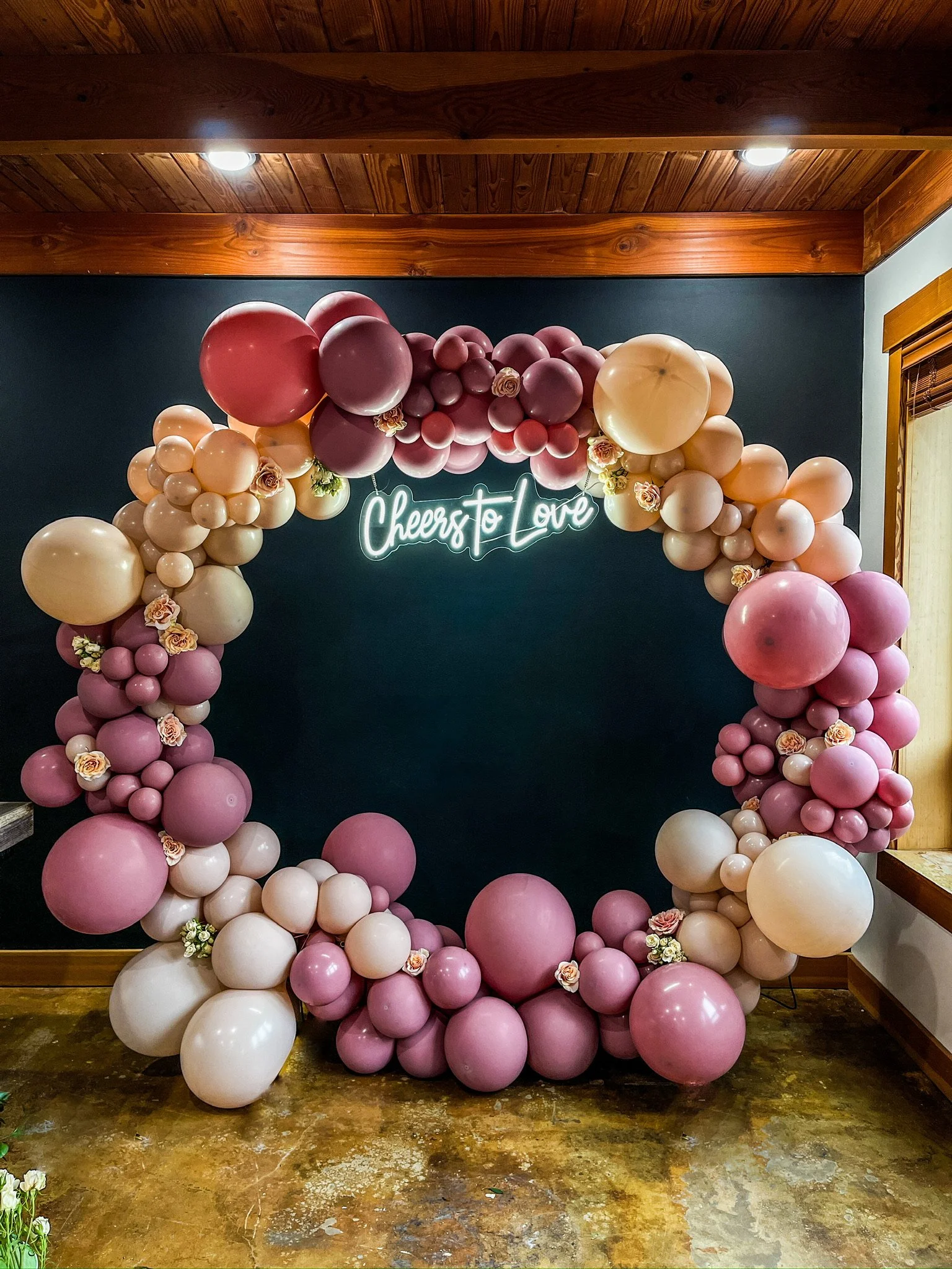 Decorative balloon arch with pink, cream, and burgundy balloons surrounding a neon sign that reads 'Cheers to Love' in a room with dark walls and wood accents.