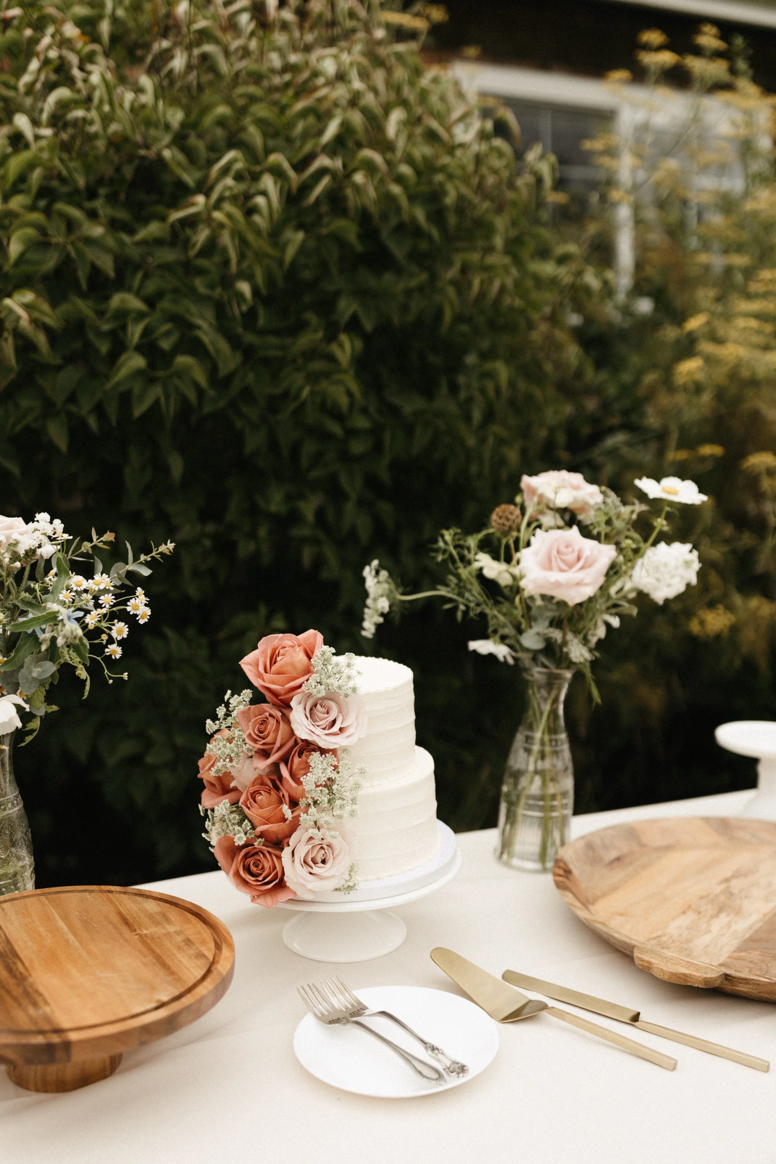 A white wedding cake decorated with pink and white roses, placed on a white table with a wooden tray, a small plate with a fork, and gold-colored cake serving utensils. There are vases with flowers and a leafy green background.