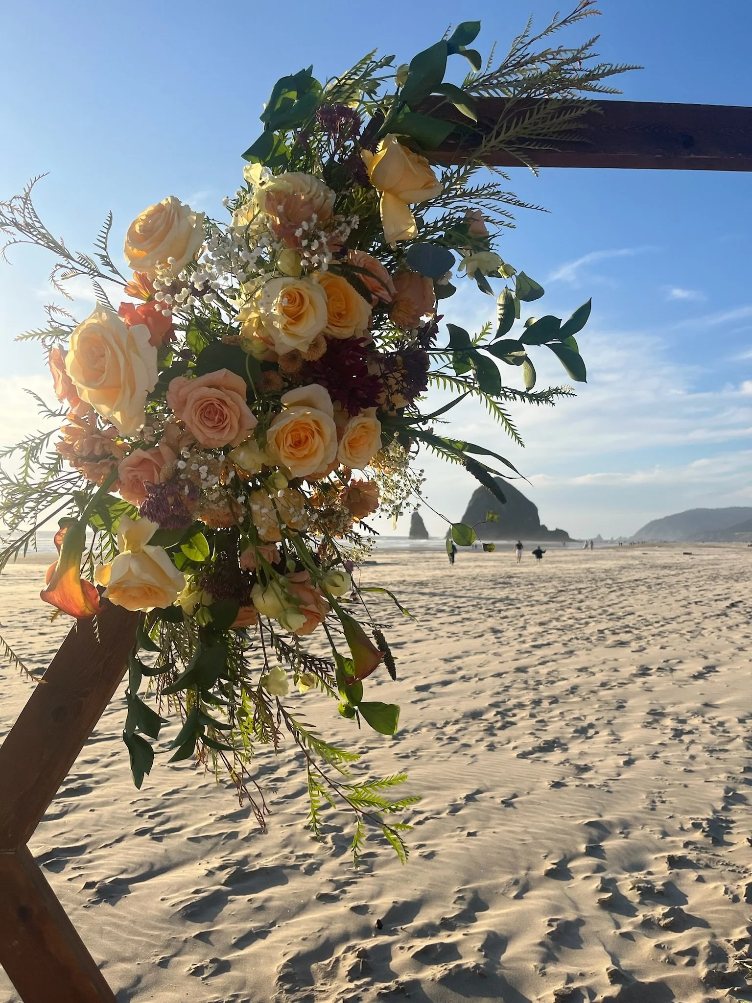 A floral arrangement of roses, greenery, and other flowers attached to a wooden structure on a sandy beach with rock formations in the background during sunset.