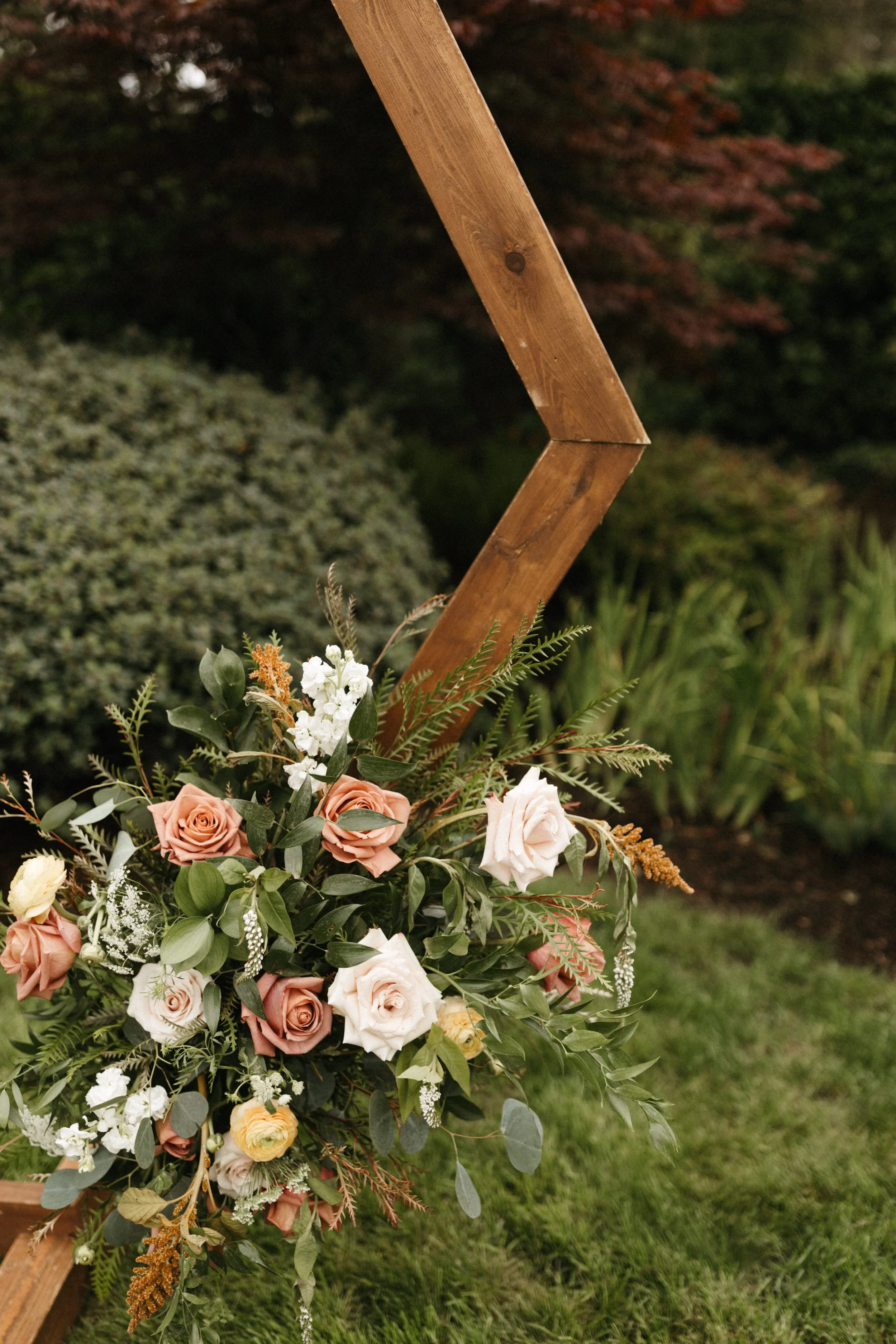 A floral arrangement featuring pink and white roses with greenery, attached to a wooden frame outdoors.