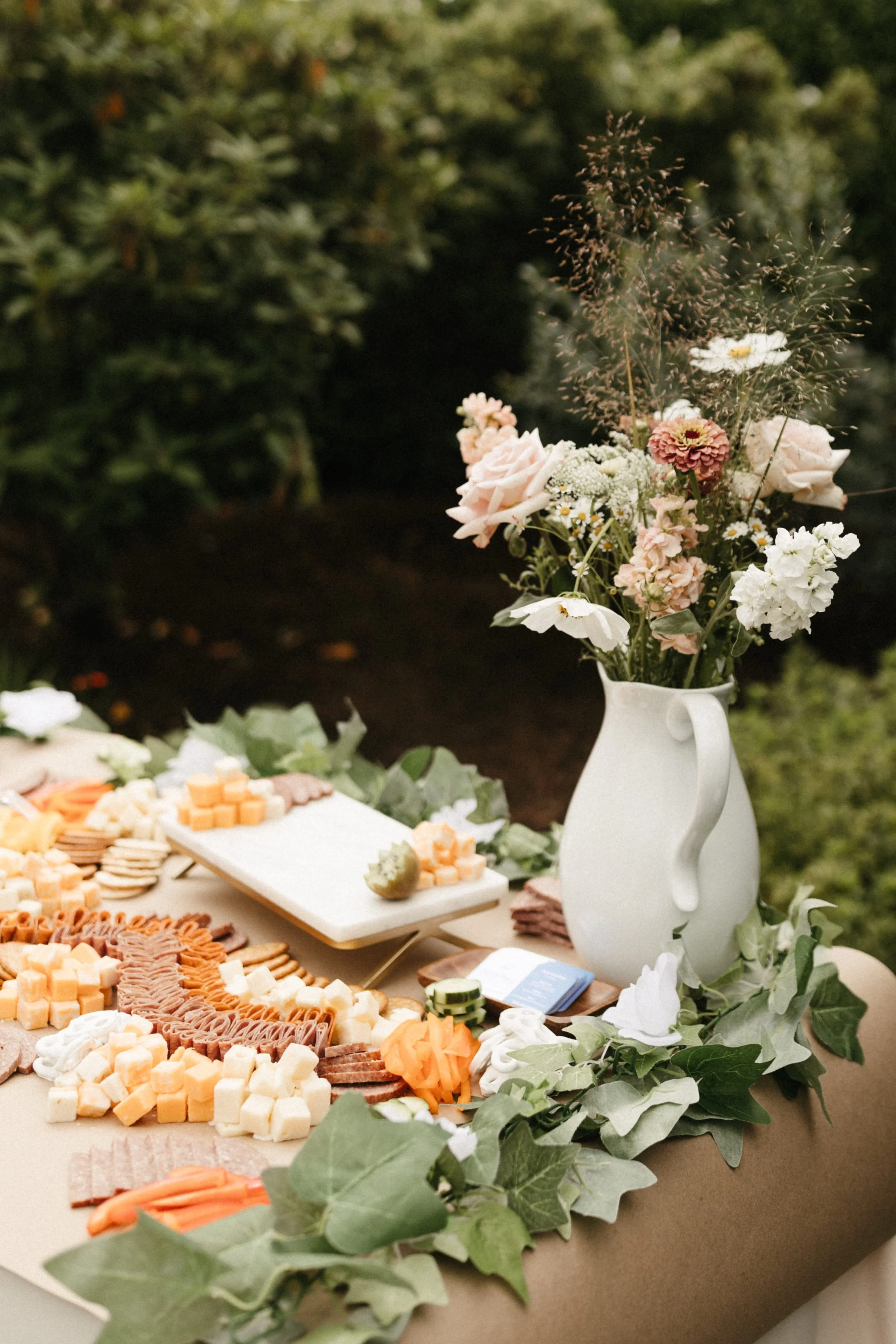 A cheese and charcuterie platter with various cheeses, sliced meats, and vegetables on a table decorated with greenery, alongside a white pitcher holding a bouquet of pink and white flowers, outdoors with a greenery background.