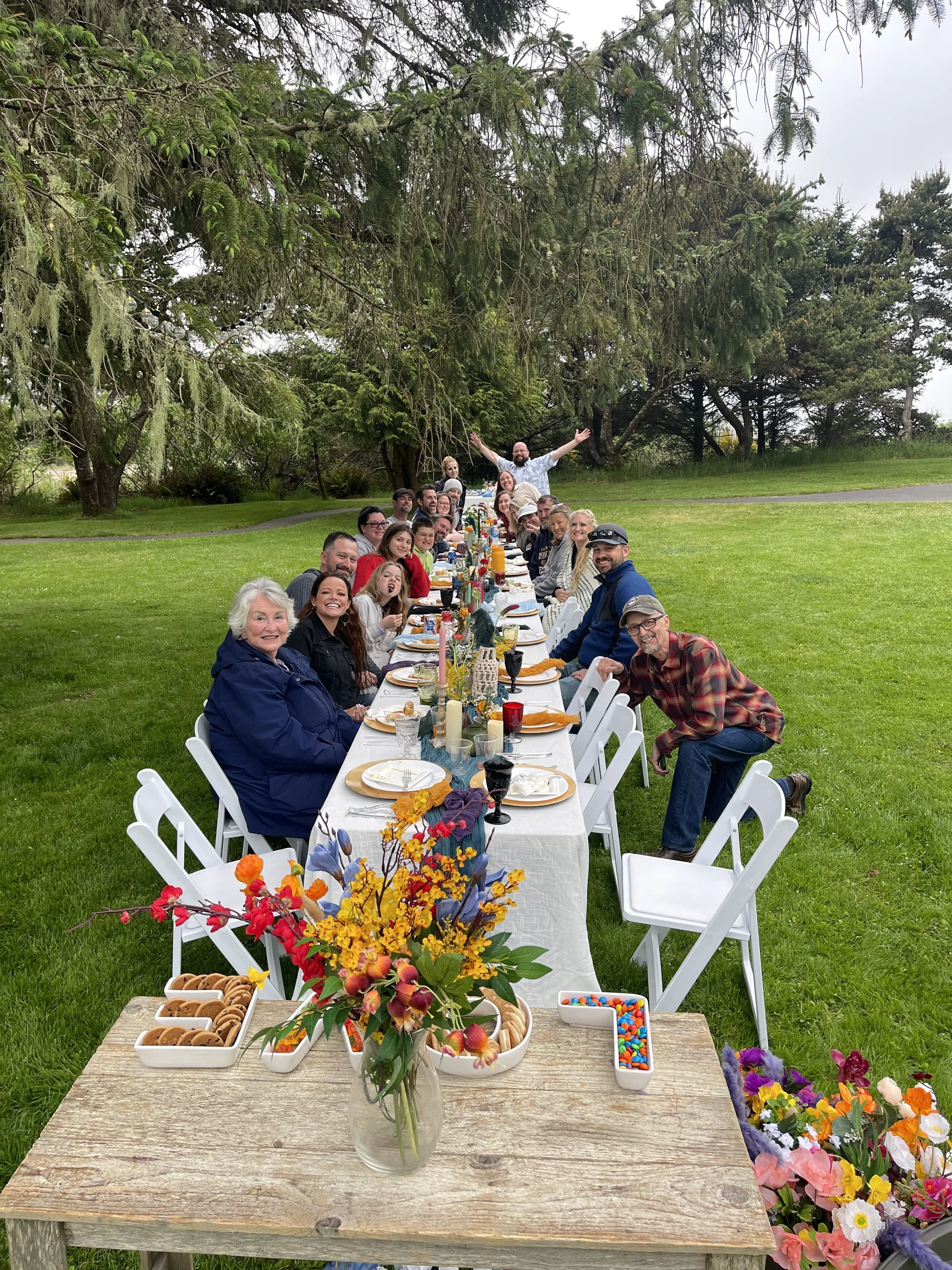Long outdoor dinner table set on a grassy lawn with people celebrating, surrounded by trees, with food, drinks, flowers, and a group of people smiling and posing for a photo.