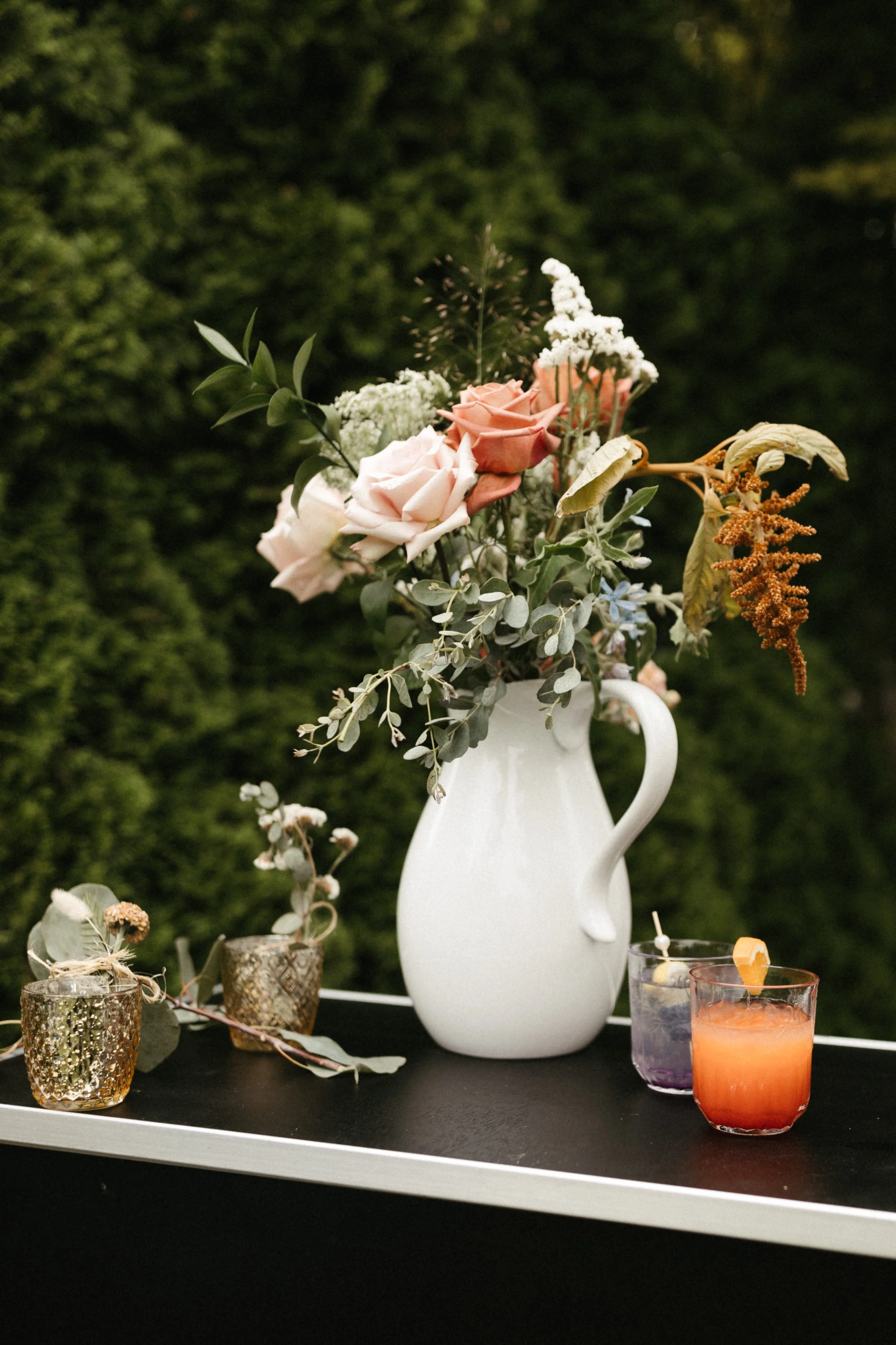 A white pitcher holding a bouquet of pink, peach, and white roses, greenery, and dried flowers on a black surface with small decorative candles and colorful drinks, against a green outdoor background.