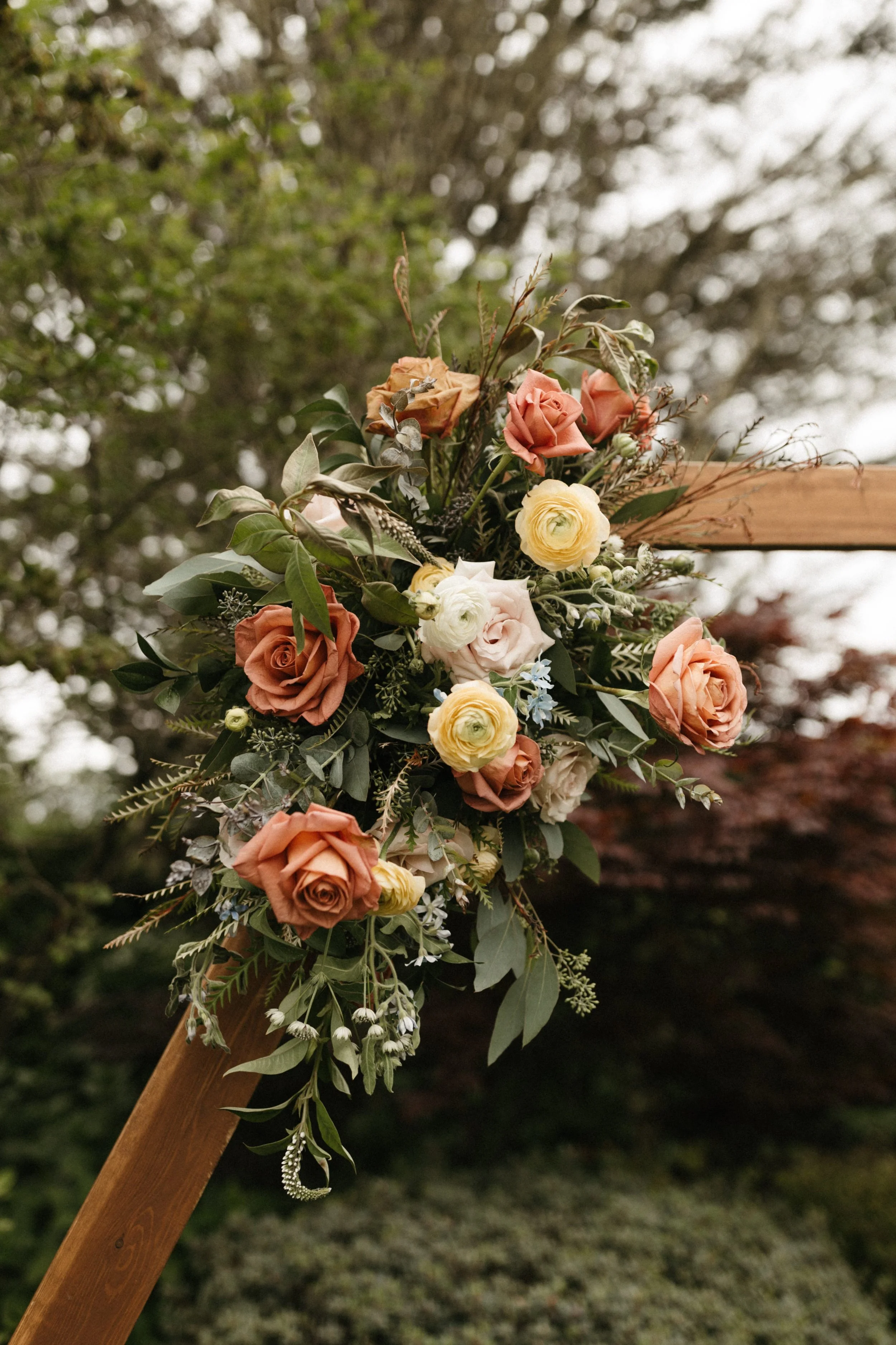 A floral arrangement with pink, peach, yellow, and white roses and greenery attached to a wooden structure outdoors.
