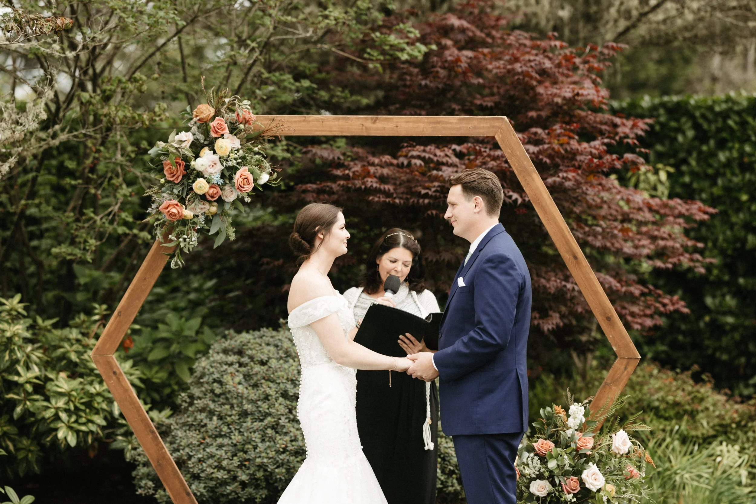 A couple exchanging vows during a wedding ceremony outdoors, with a floral arch and a woman officiant holding a microphone.