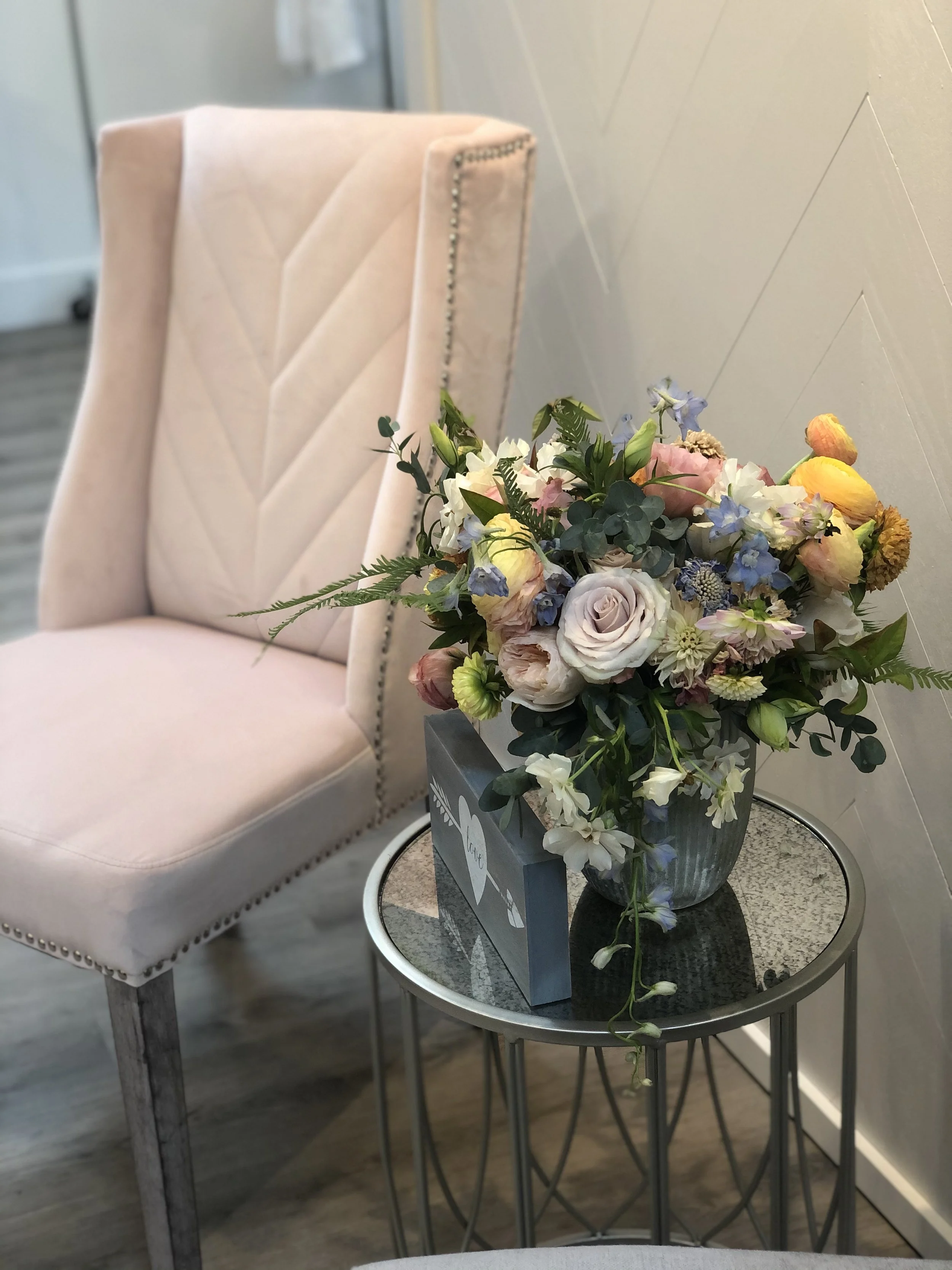 A pink upholstered armchair with nailhead trim, placed next to a round glass-top side table holding a flower arrangement in a striped vase and a boxed sign with flower illustrations.