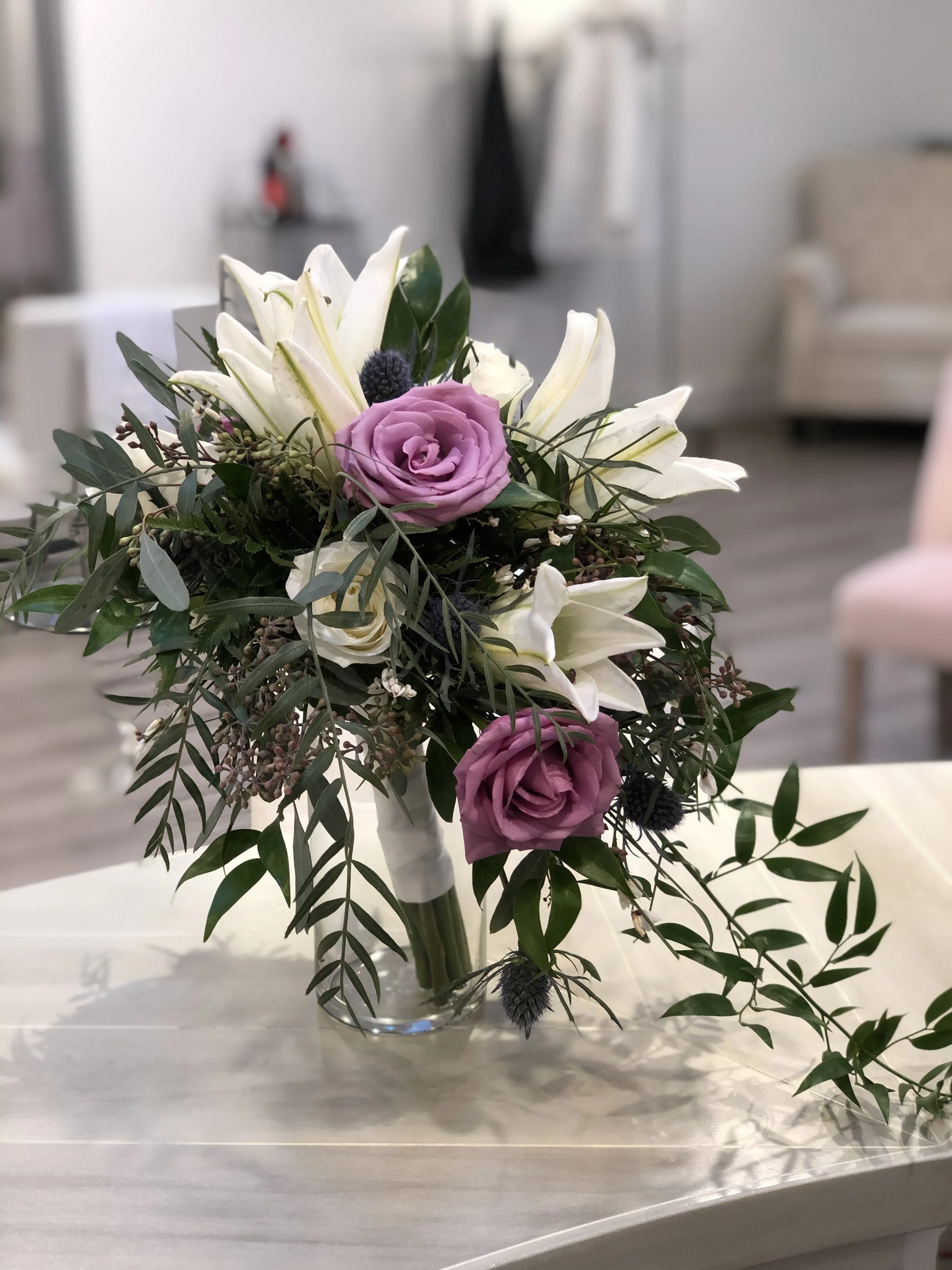 Flower arrangement with white lilies, purple roses, and greenery in a clear glass vase.