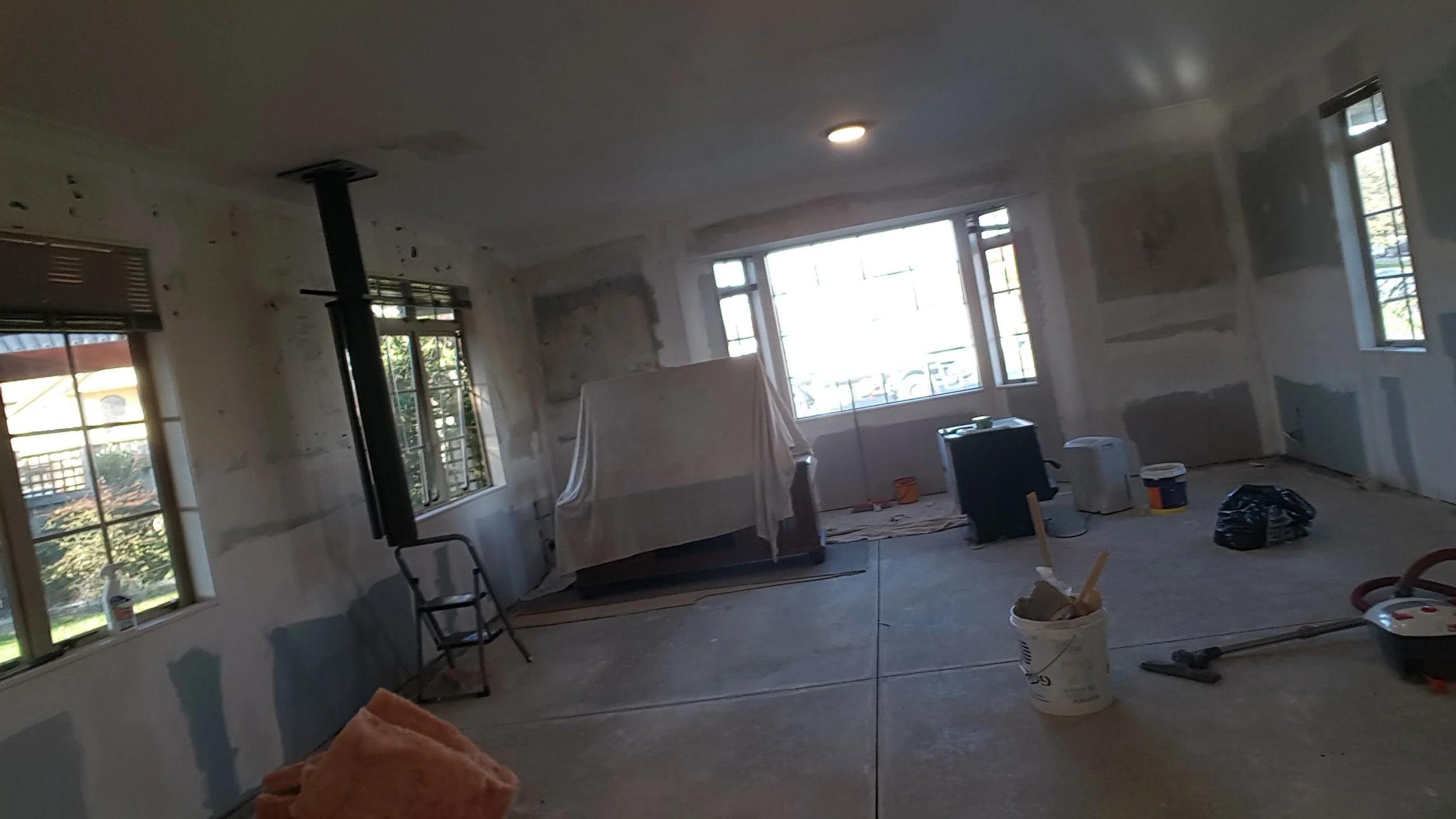 Room undergoing renovation with drywall patches, construction tools, and covered furniture, illuminated by natural light from multiple windows.