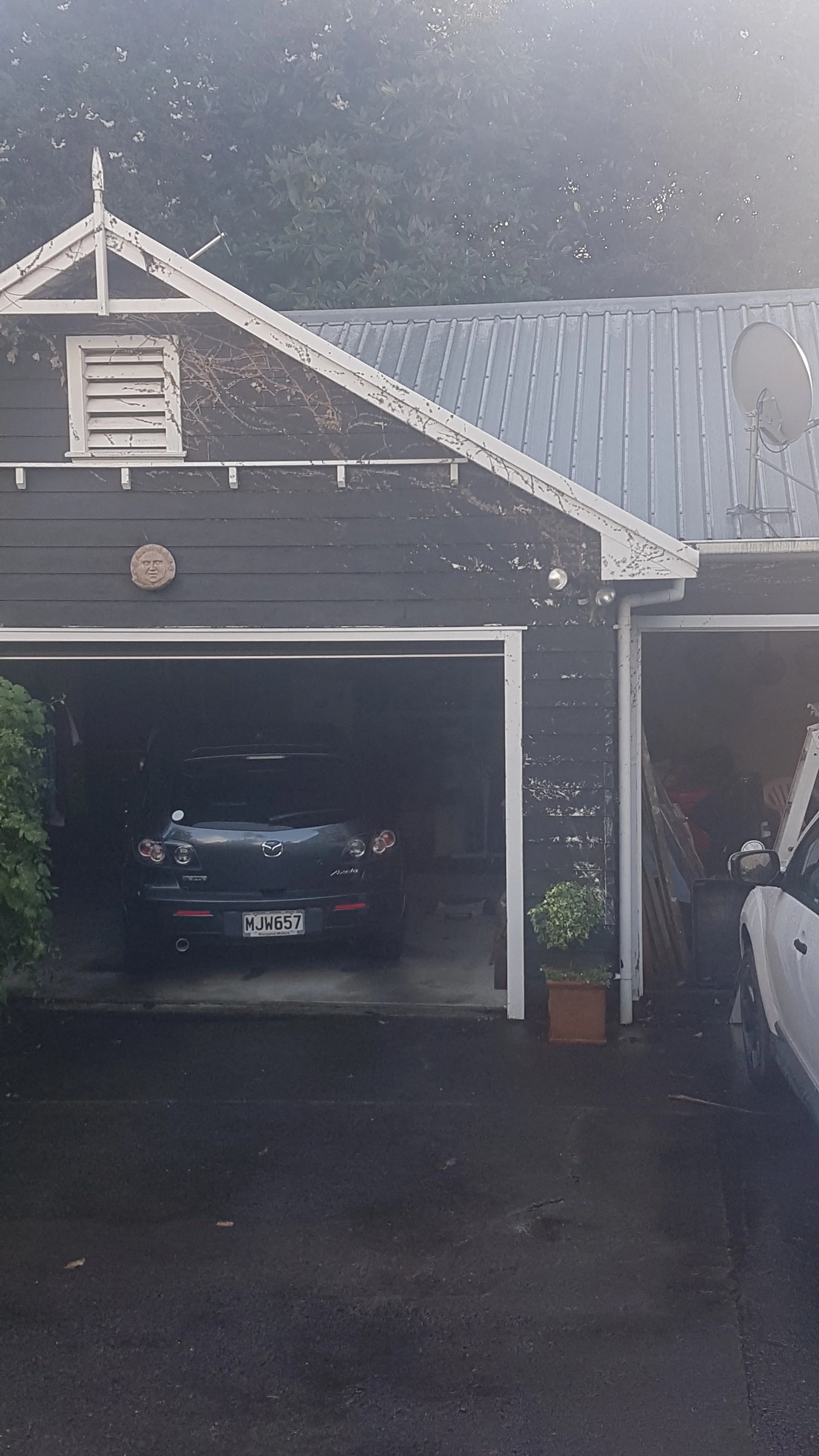 A house with dark siding, a sloped roof, and a small window with white shutters. A satellite dish is mounted on the roof. A black Mazda car is parked in the garage, and a white vehicle is partially visible beside it. There is a decorative face on the