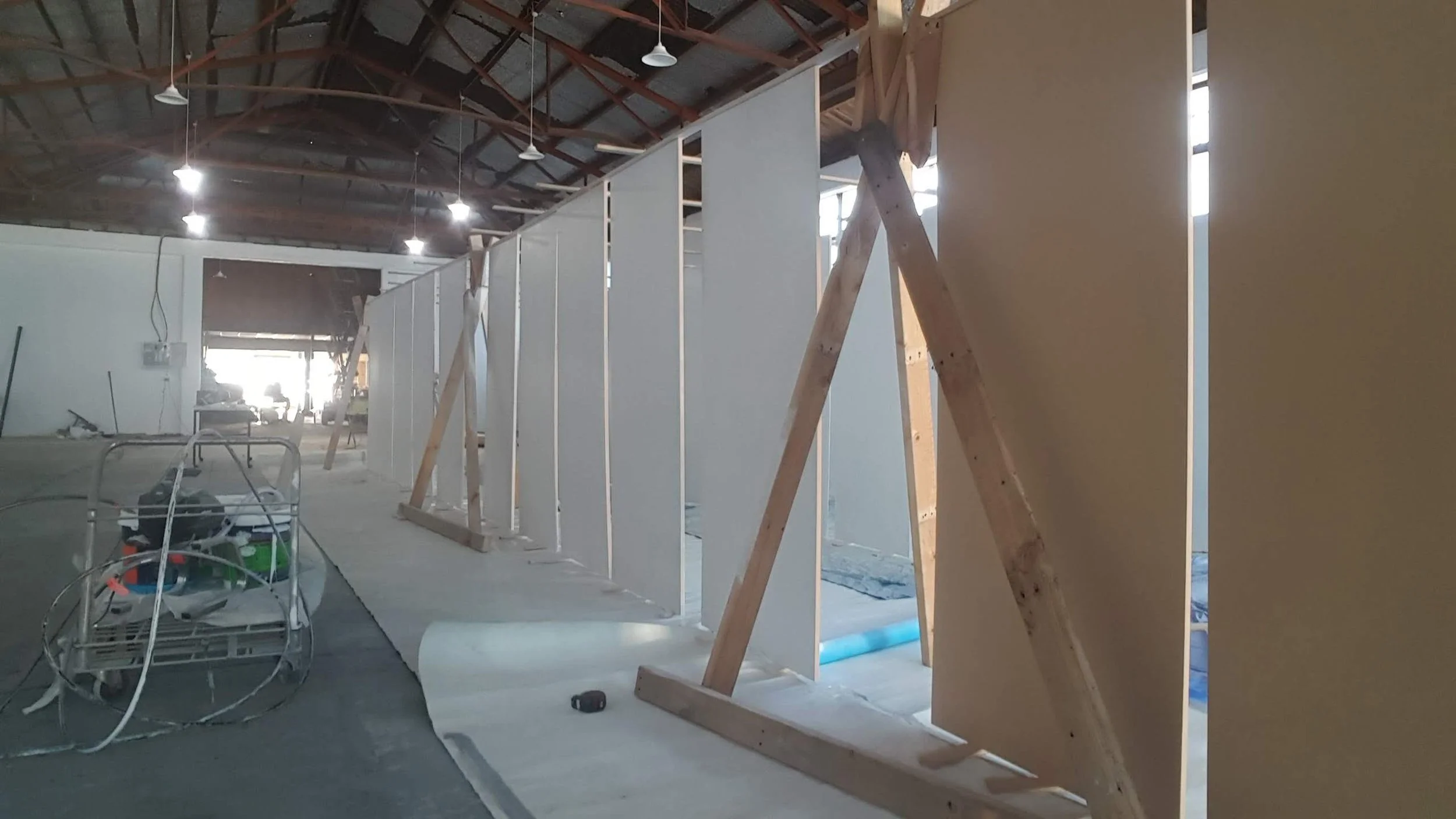 Construction inside a warehouse showing partially built partition walls with wooden framework and white panels, construction tools and equipment on the floor.
