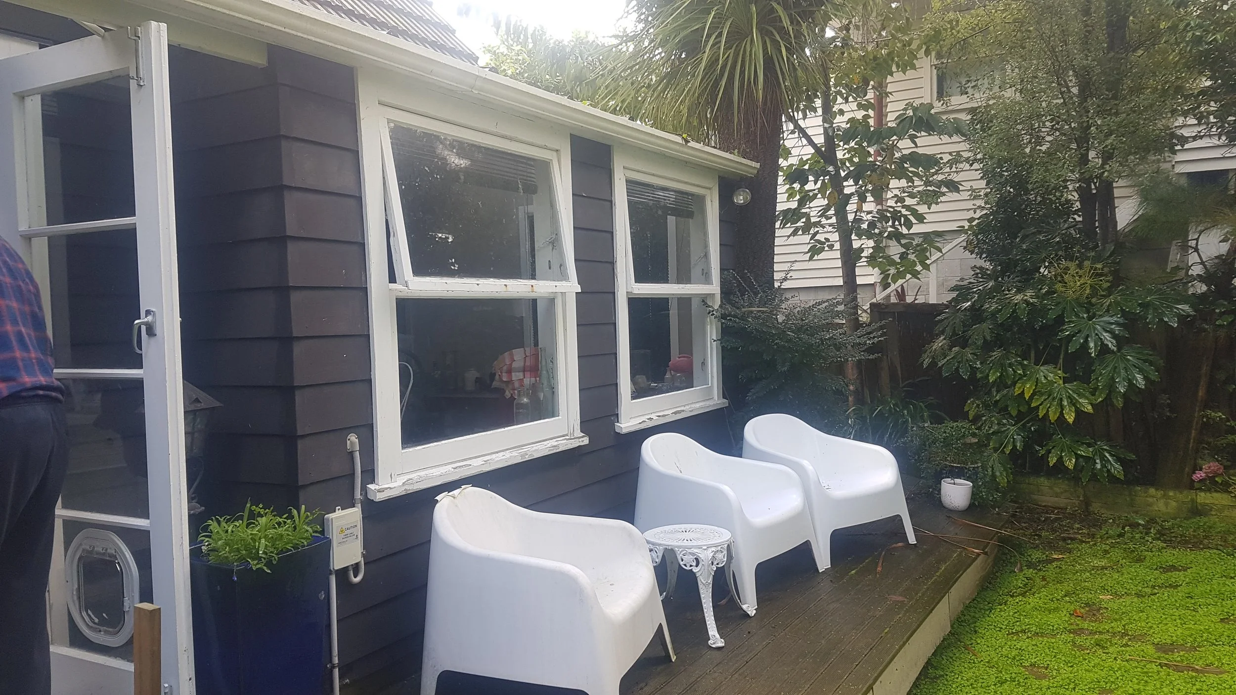 Backyard patio with three white plastic chairs, a small ornate white table, and lush greenery surrounding a blue house with three large windows.