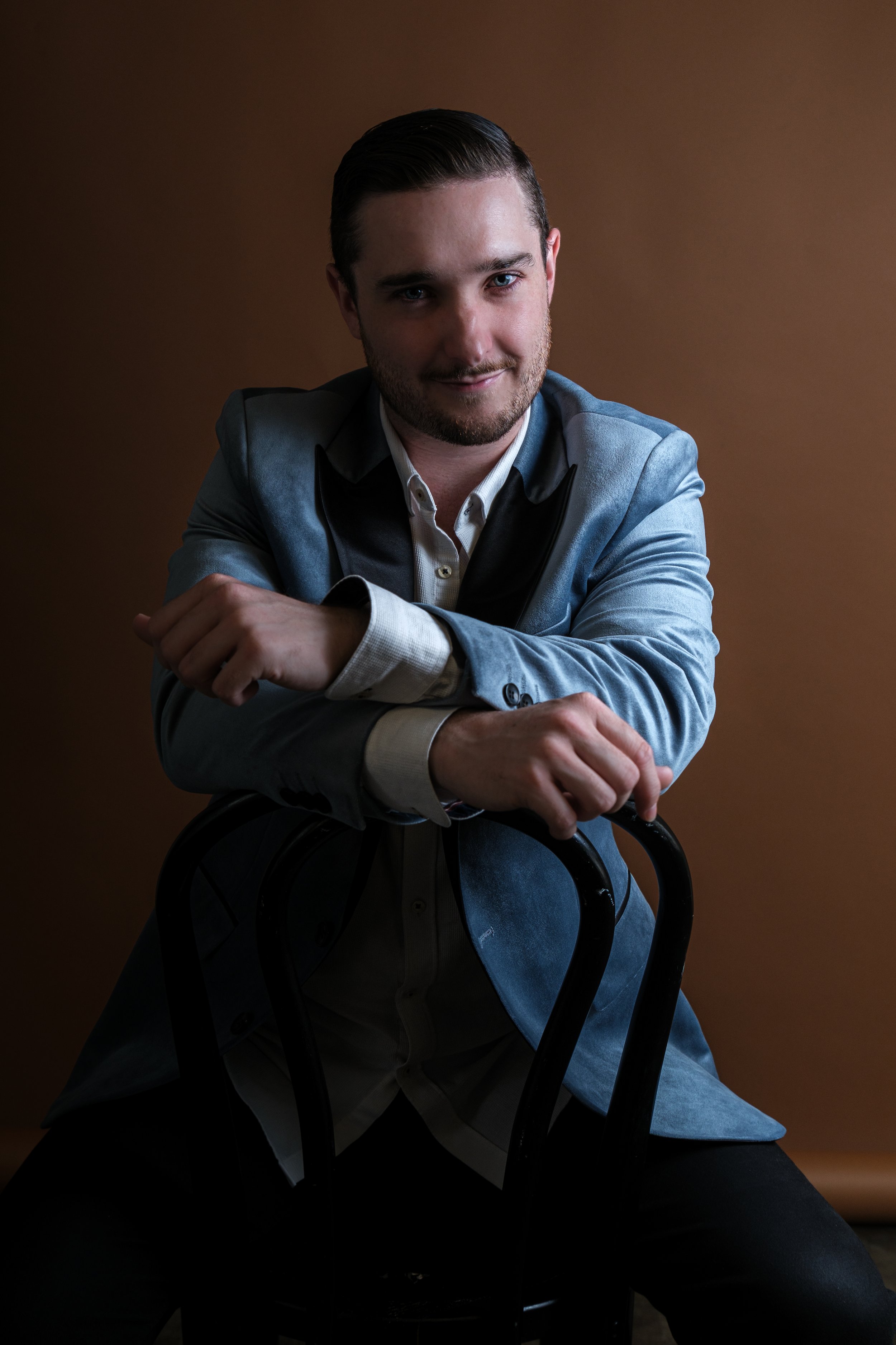 A man with dark hair and a beard, wearing a blue blazer over a white shirt, sits on a black chair against a brown background, looking at the camera with a slight smile.