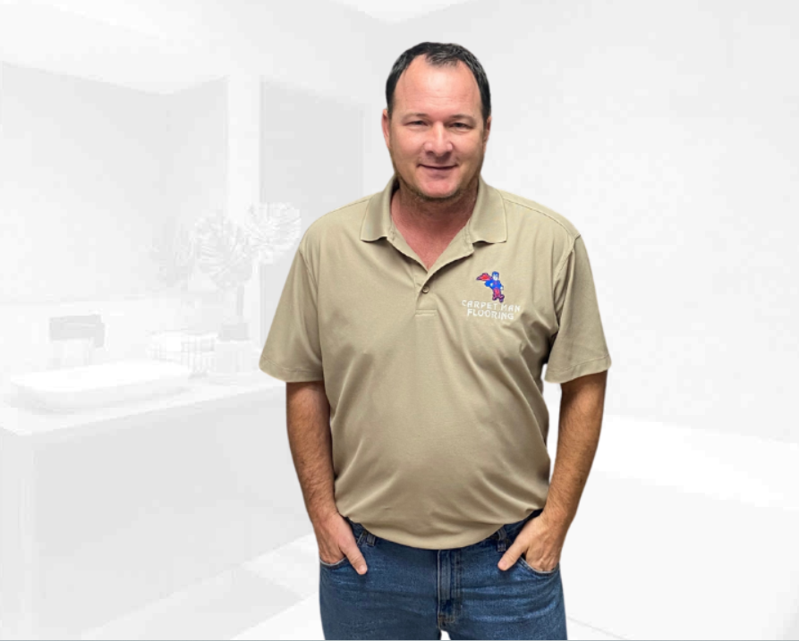 Man in beige polo shirt with Carpets and Flooring logo, standing indoors with hands in pockets, smiling.